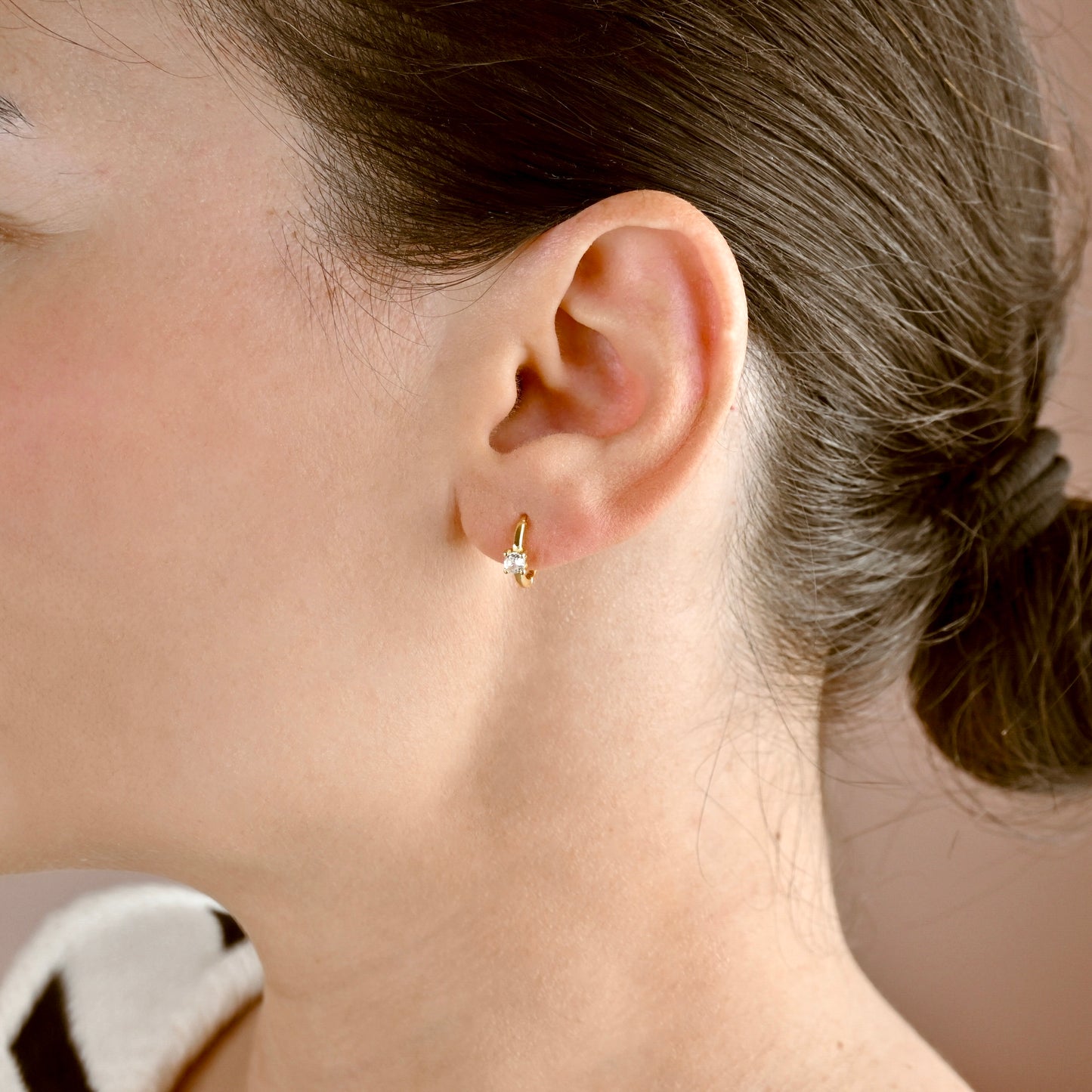 A close-up of a woman's ear wearing a small gold huggie hoop earring with a tiny clear gemstone, shown against her skin and hair.