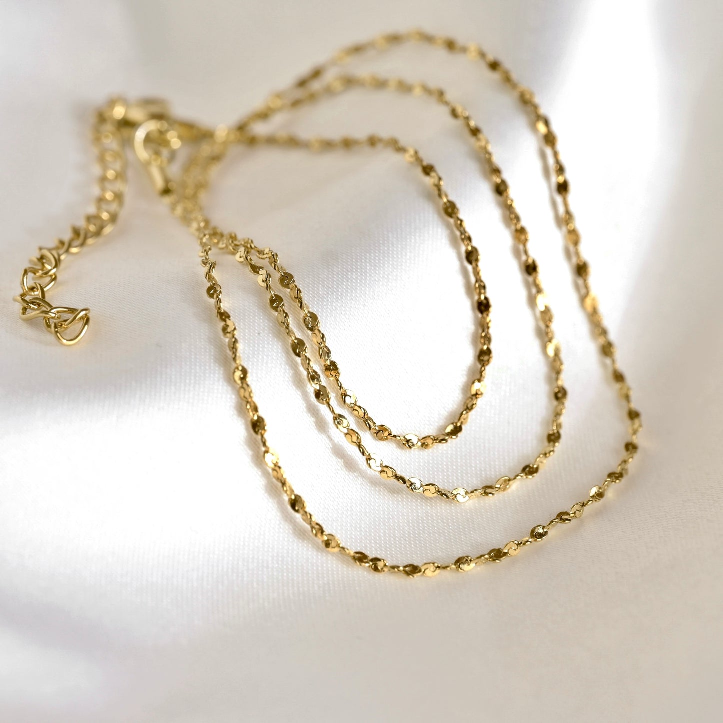 A delicate gold chain necklace with a textured, faceted link design, coiled on a white satin fabric background.