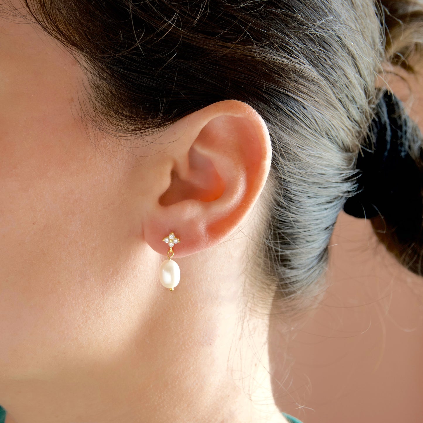 A close-up of a person's ear wearing a gold stud earring with a small floral cluster and a dangling white pearl.