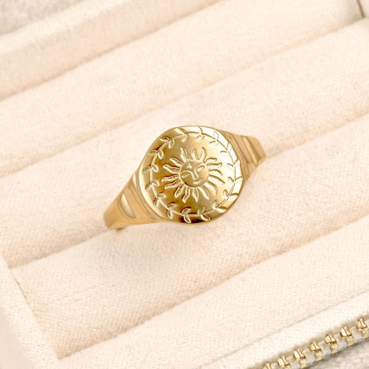 A gold signet ring featuring an engraved  ancient sun motif and leafy wreath design, displayed on a beige textured jewelry box.