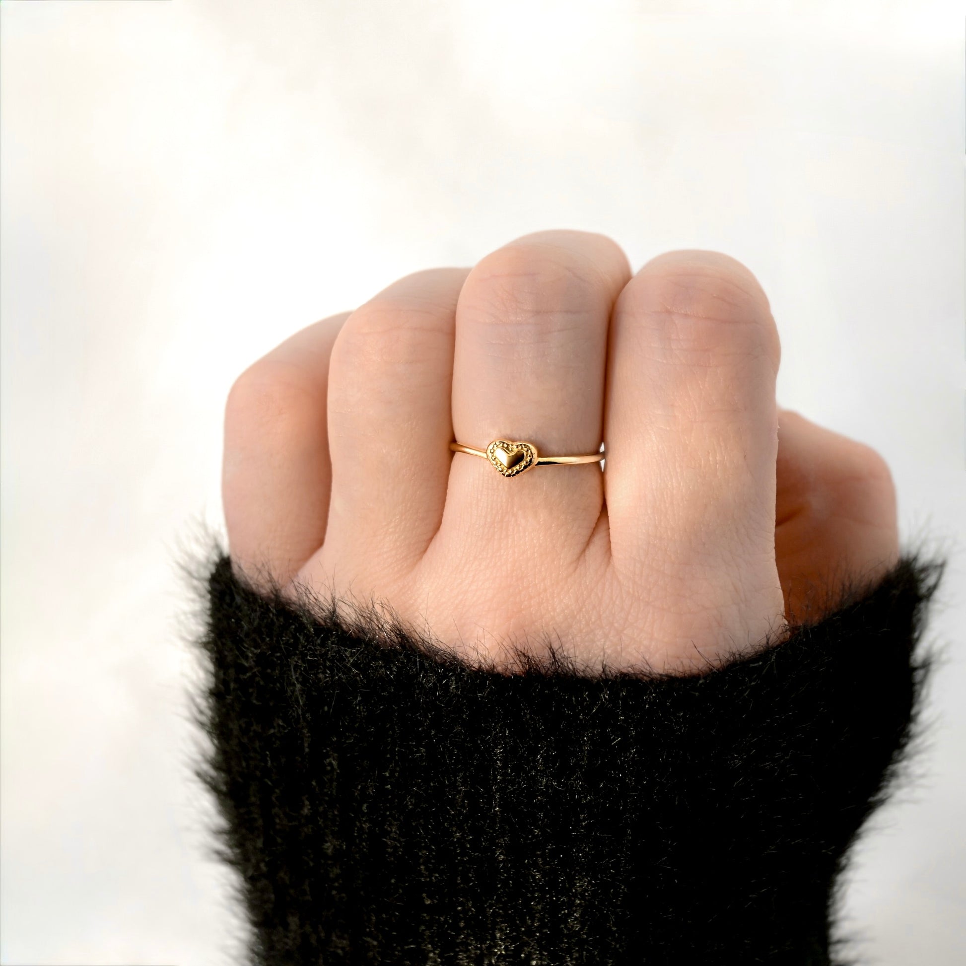 A close-up of a hand wearing a delicate gold ring with a small heart-shaped charm, set against a soft, out-of-focus light background.