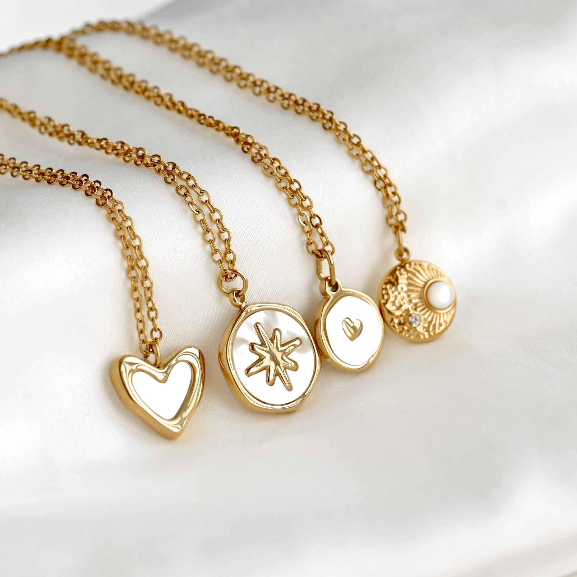 Four gold chain necklaces with white inlay pendants are arranged on a soft white fabric background. The pendants include a heart shape, a starburst design, a small heart, and a textured sunburst with a pearl accent.