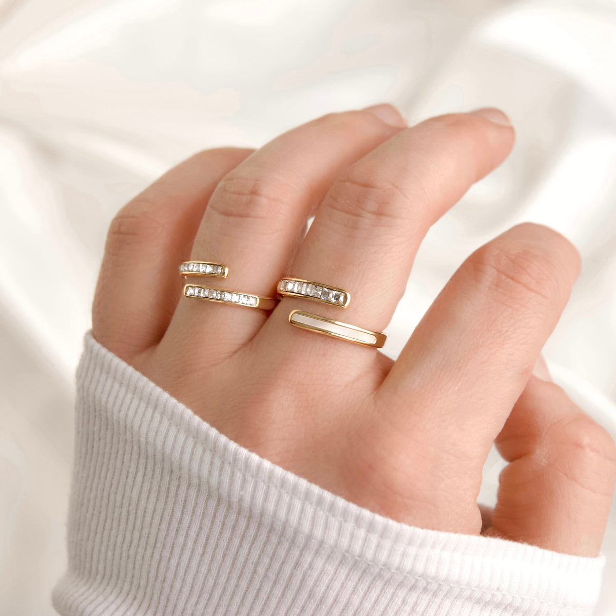 A close-up of a hand wearing two bypass gold rings, including one with small clear stones, and one with shell inlay and cz, against a soft white fabric background.