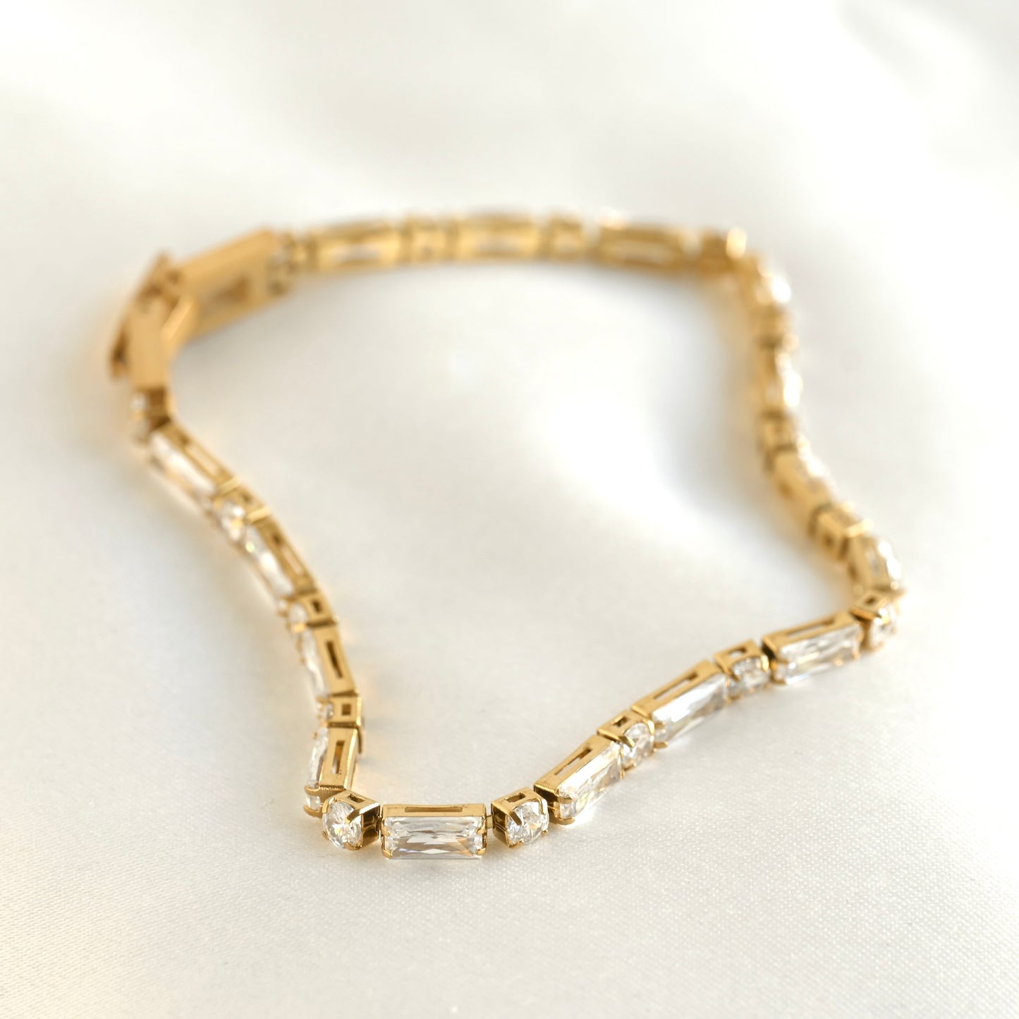 A gold-plated tennis bracelet featuring baguette-cut clear crystals, displayed on a soft white satin fabric.