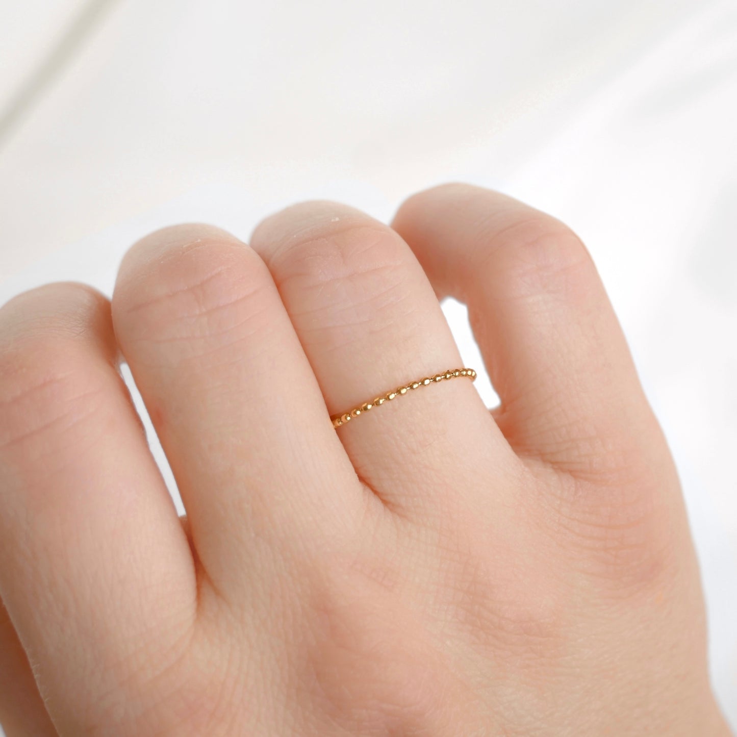 A close-up of a hand wearing a delicate, thin gold beaded chain ring on one of the fingers, set against a soft, light-colored fabric background.