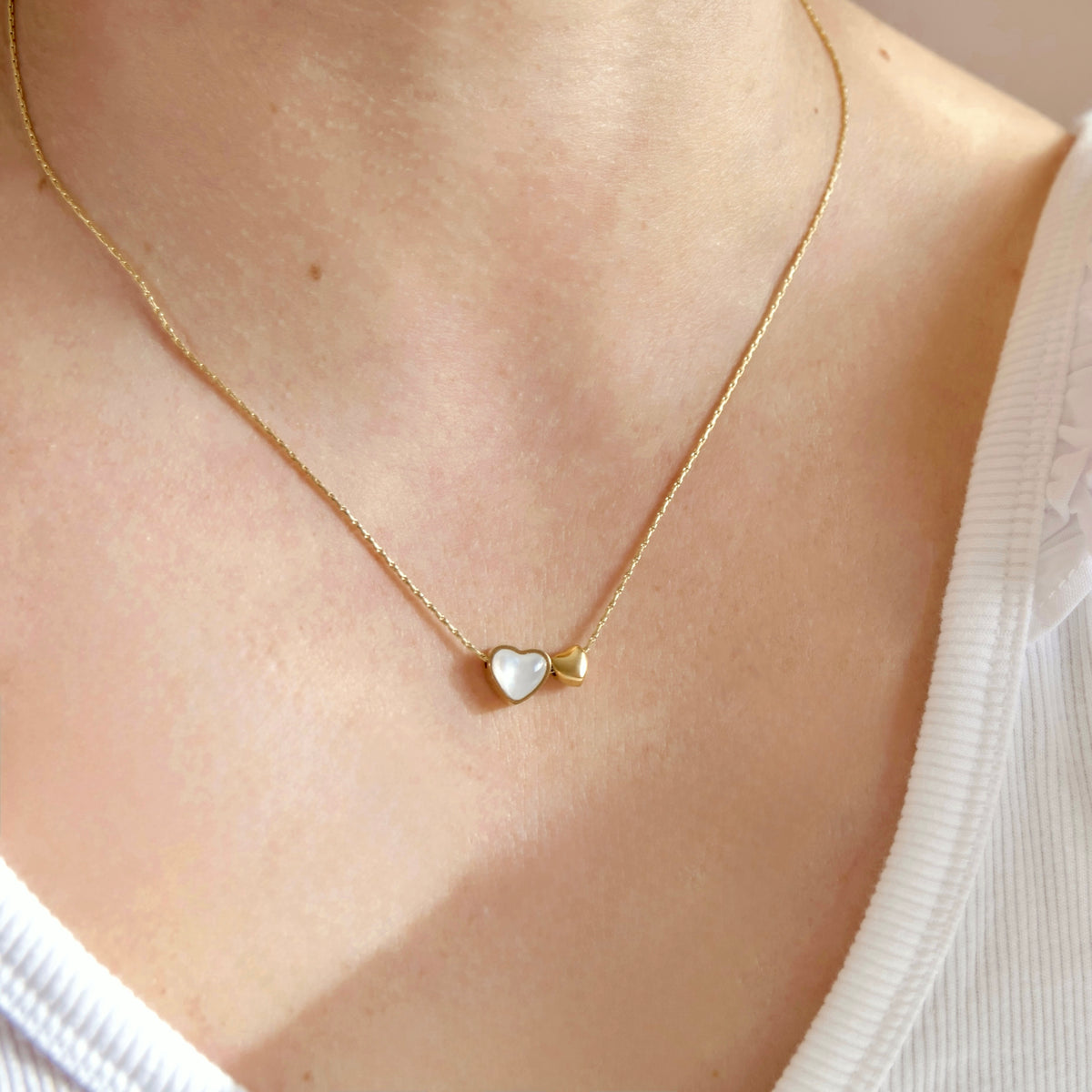 A close-up of a person's neck and upper chest wearing a delicate gold chain necklace with a small double-heart pendant, one heart in white and one in gold, over a white ribbed top.