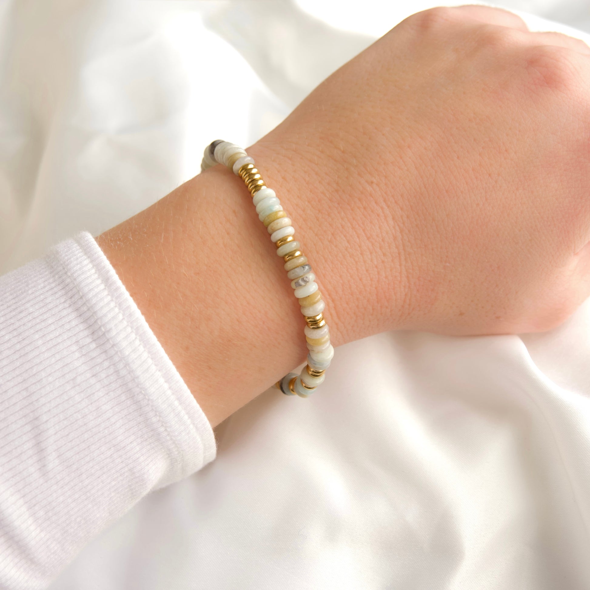 A close-up of a person's wrist wearing a beaded bracelet made of light green and white stone beads with small gold spacer accents, set against a soft white fabric background.