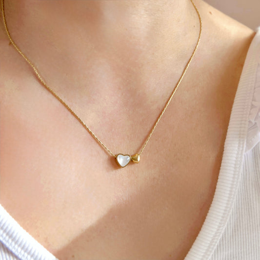 A delicate gold chain necklace with two small heart charms, one white and one gold, worn on a person's neck over a white ribbed top.