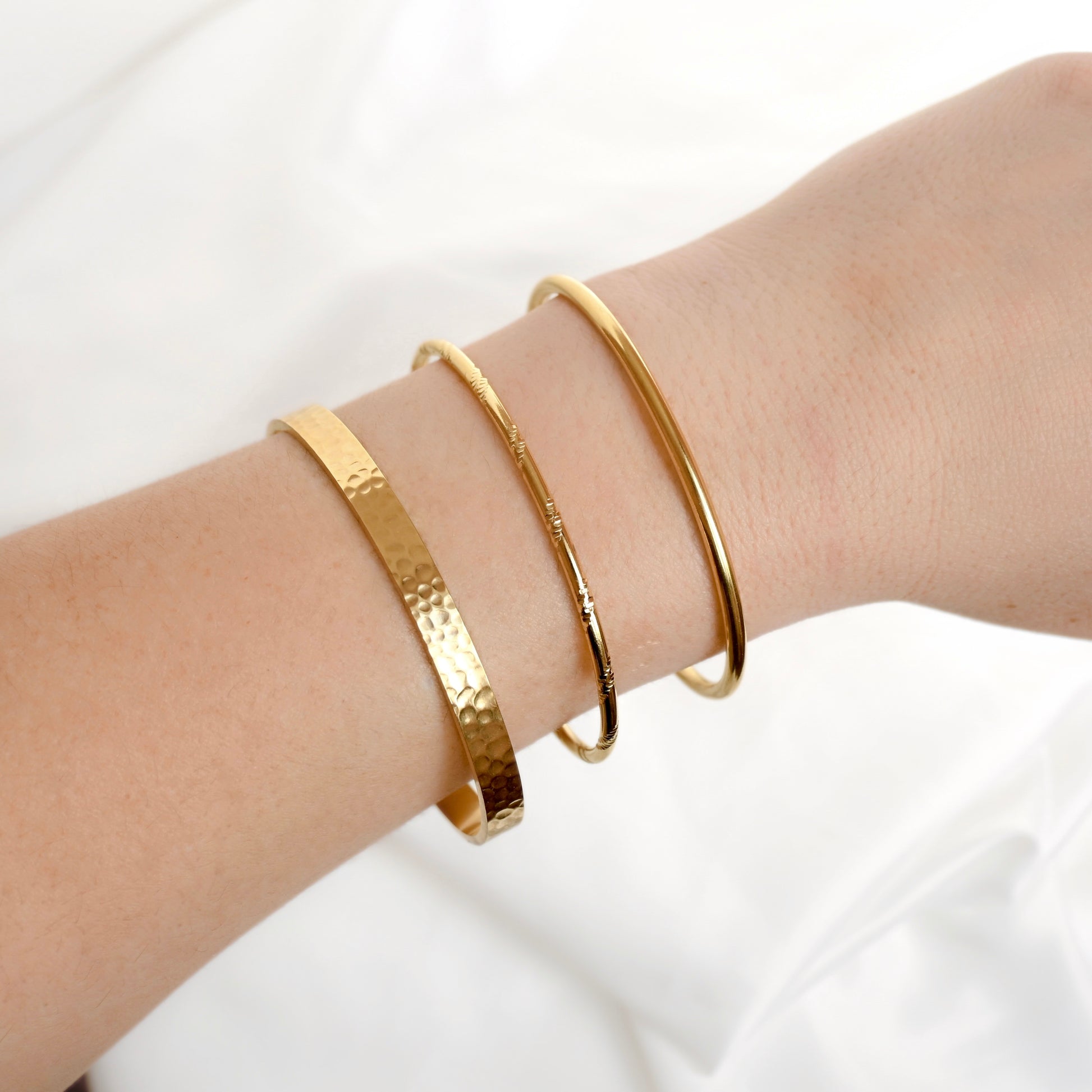 Three gold bangle bracelets of different textures and designs worn on a person's wrist against a soft white fabric background.