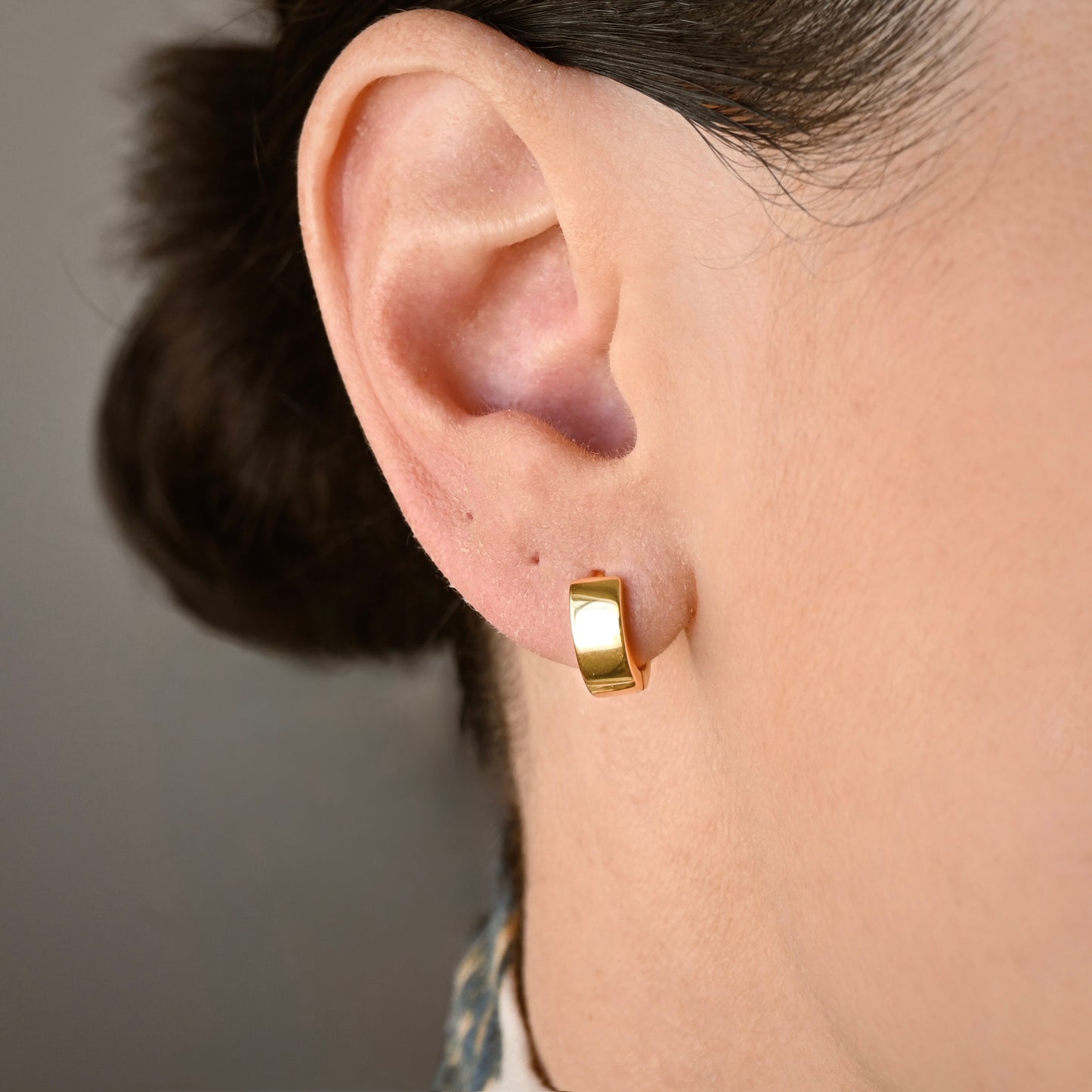 A close-up of a person's ear wearing  small thick gold huggie hoop earrings against a soft gray background.