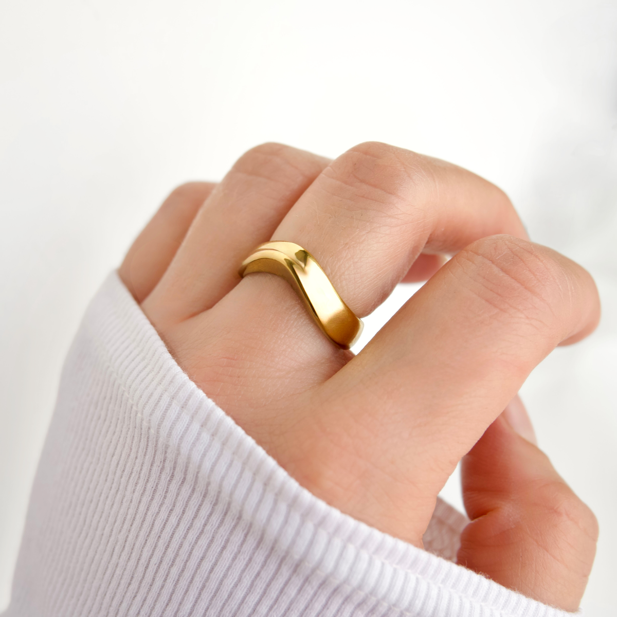 A close-up of a hand wearing a polished gold band ring with a wavy, curved profile, set against a clean white background.
