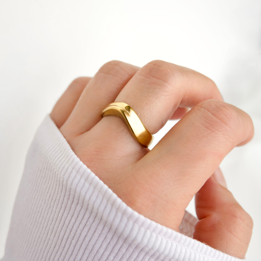A close-up of a hand wearing a polished gold band ring with a wavy, curved profile, set against a clean white background.