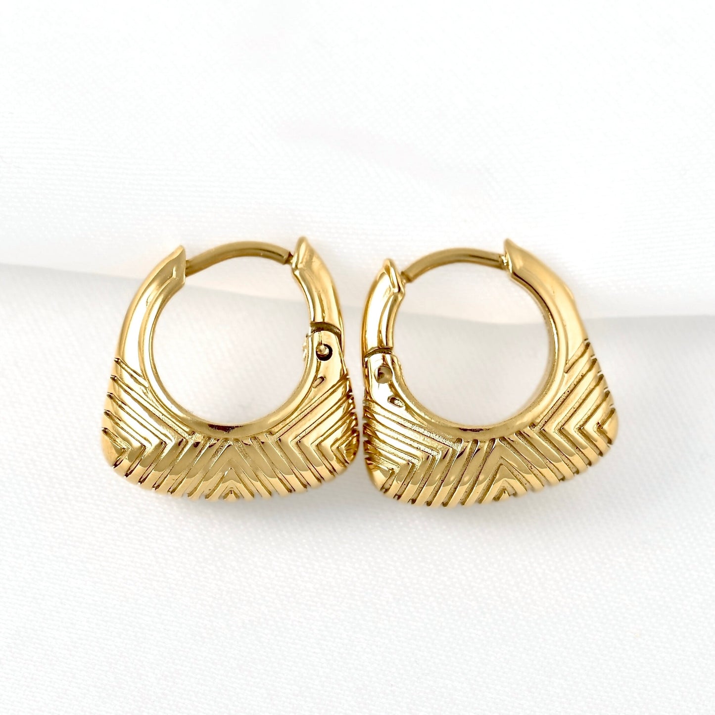 A pair of gold huggie hoop earrings with a textured chevron pattern on the front, displayed on a clean white background.