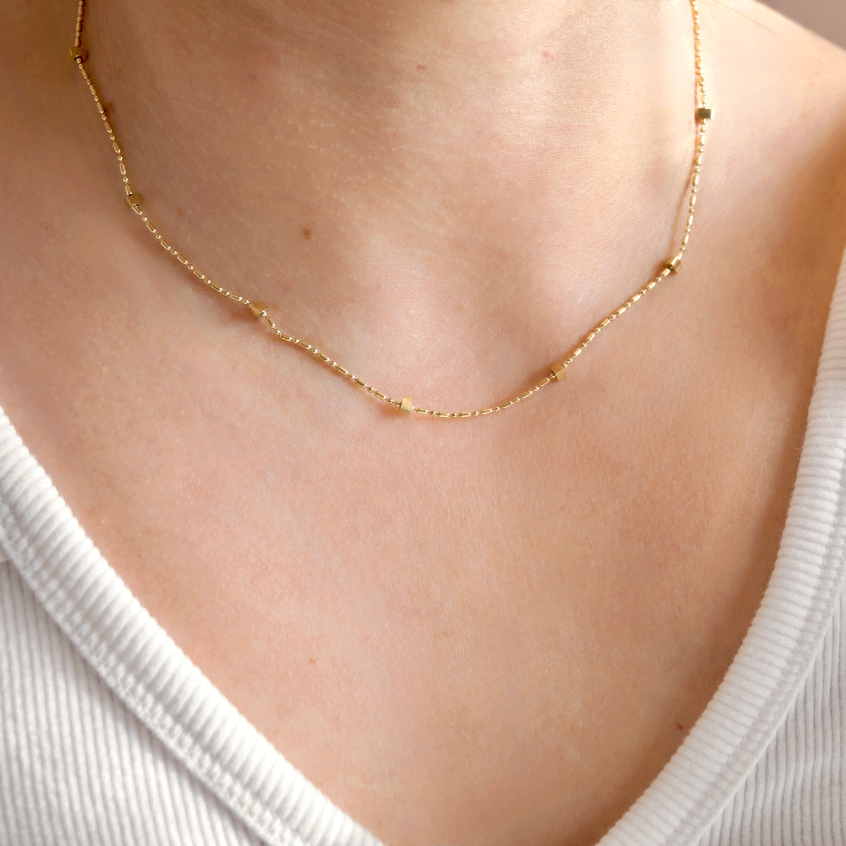 A delicate gold chain necklace with small geometric bead accents, worn on a person's neck over a white ribbed top.