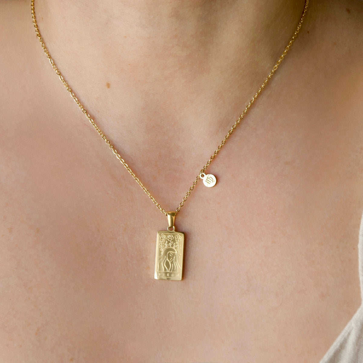 A gold chain necklace with a rectangular zodiac pendant and a small round disc charm, worn on a person's neck.