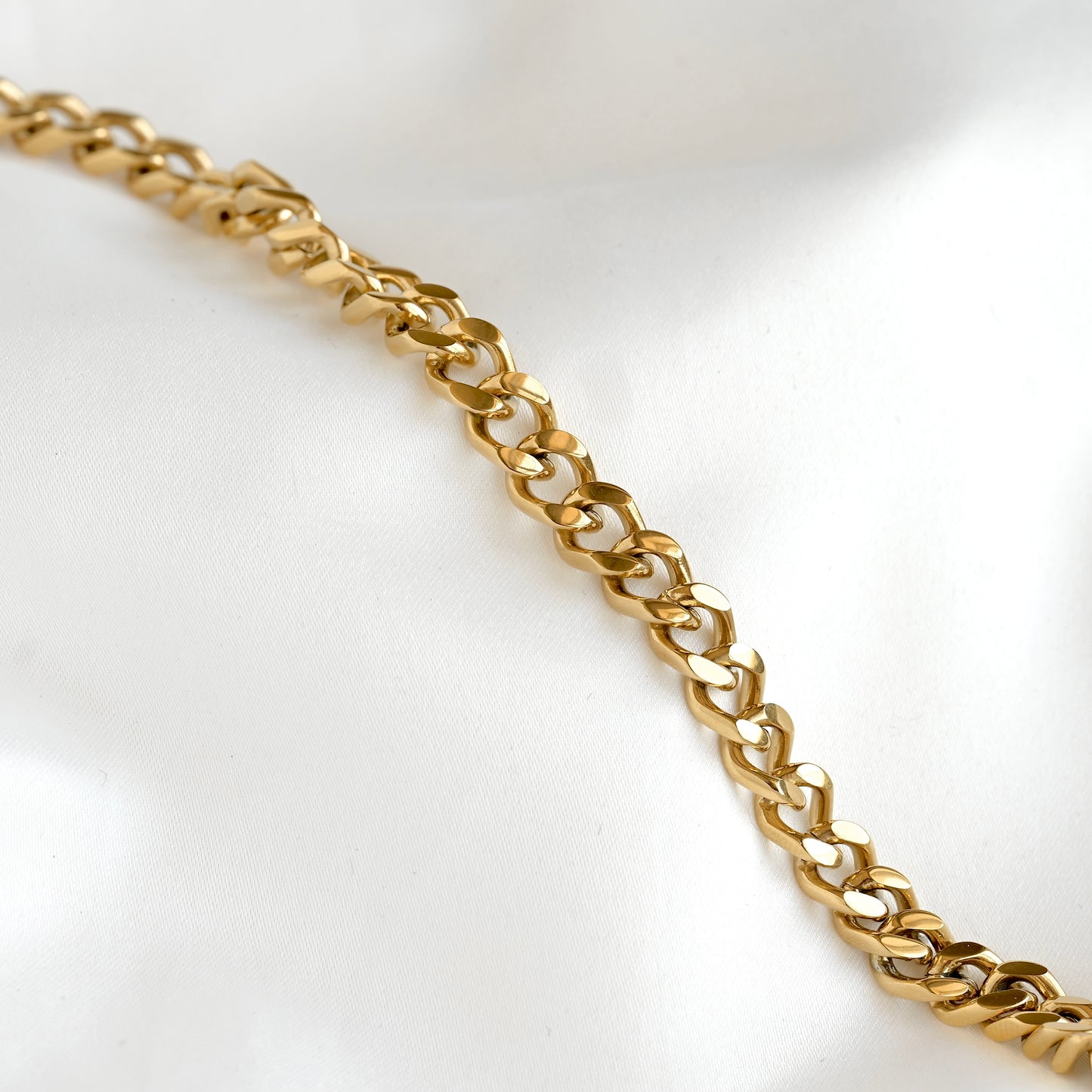 A close-up of a polished gold chain with interlocking curb-style links, laid diagonally across a soft white fabric background.