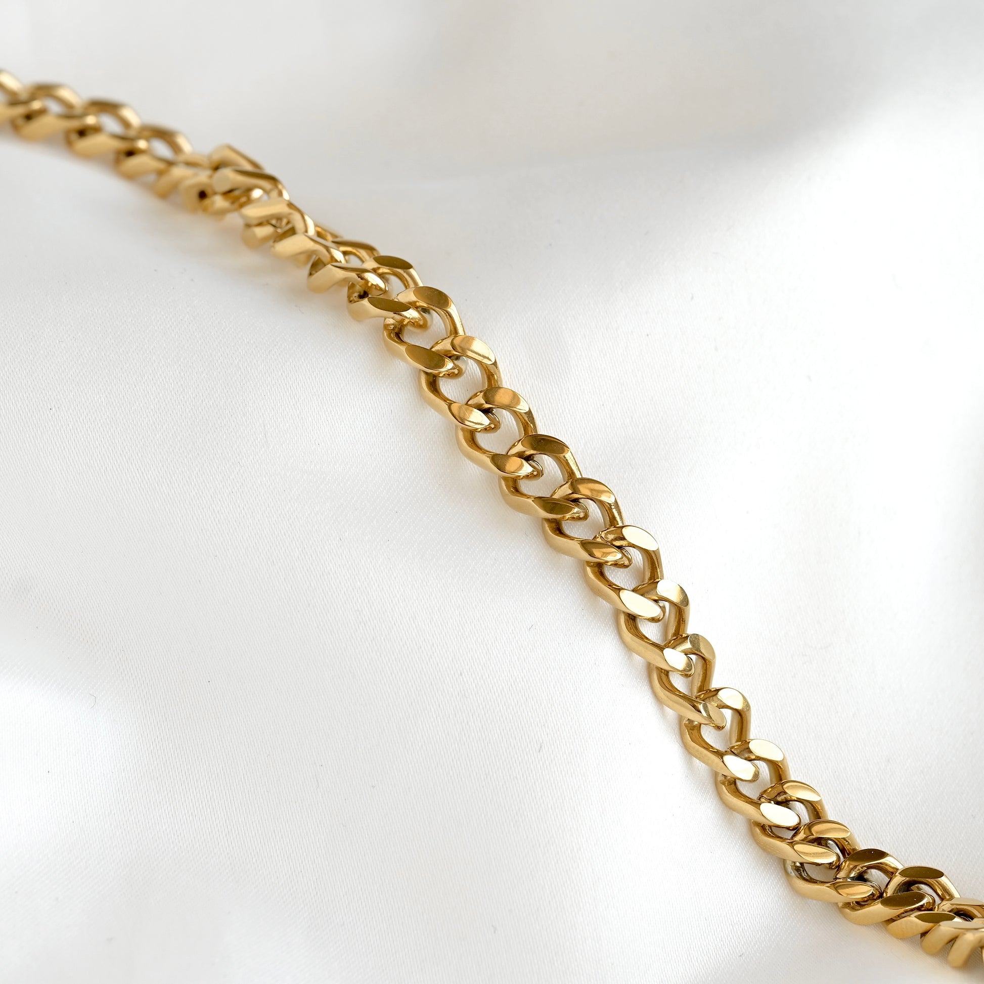 A close-up of a polished gold chain with interlocking curb-style links, laid diagonally across a soft white fabric background.
