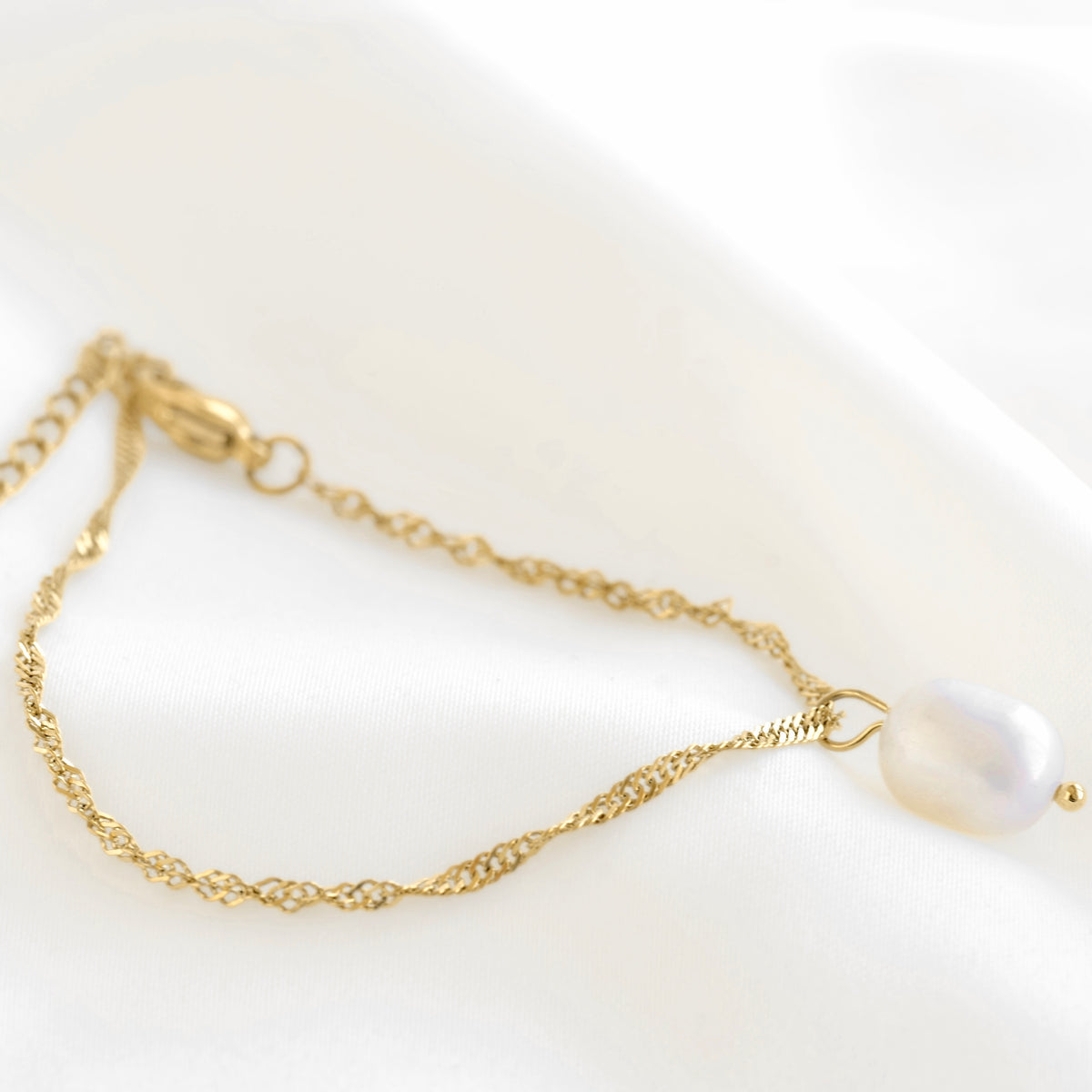 A delicate gold chain bracelet featuring a single irregular white freshwater pearl pendant, displayed on a soft white fabric background.
