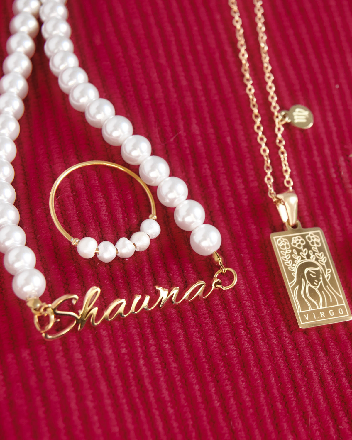 A gold and white pearl jewelry set featuring a nameplate necklace spelling 'Shauna', a matching pearl hoop, and a gold pendant with a tarot card design and 'VIRGO' text, displayed on a textured red cord background.