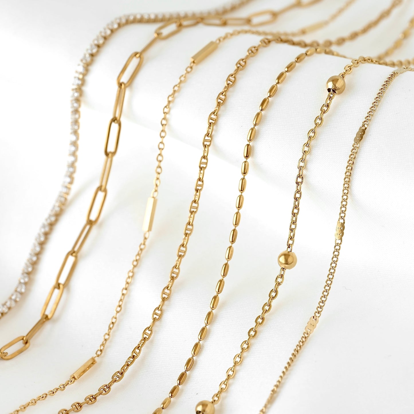 A close-up arrangement of multiple gold-tone chain necklaces in different link styles, including paperclip, curb, and tennis chains, displayed on a clean white background.