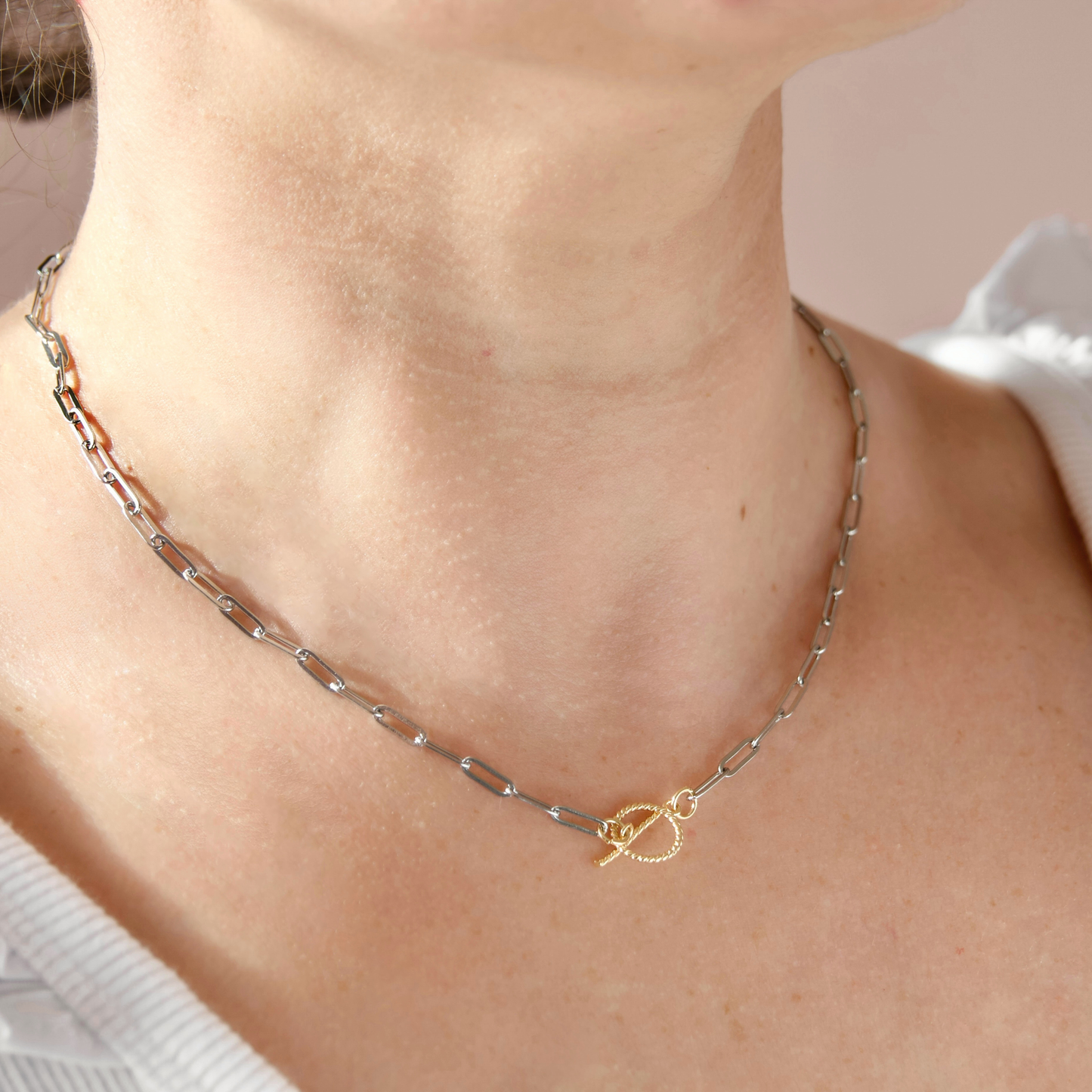 A close-up of a person's neck and upper chest wearing a delicate  paperclip chain necklace with a small twisted gold toggle clasp 