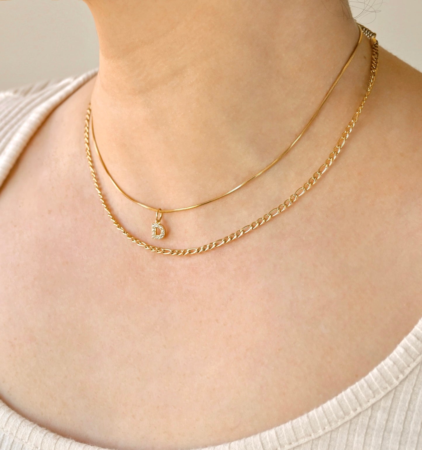 Two gold chain necklaces, one longer figaro and one shorter snake chain with diamond letter charm, on a person wearing a white top against a neutral background