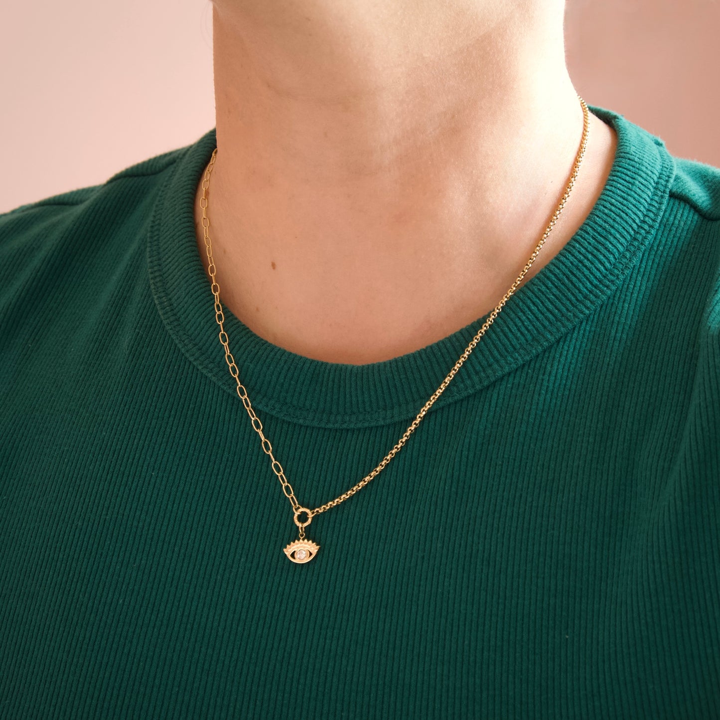 A close-up of a person's neck and upper chest wearing a delicate gold chain necklace with a small evil eye pendant, paired with a dark green ribbed crewneck shirt against a soft pink background.