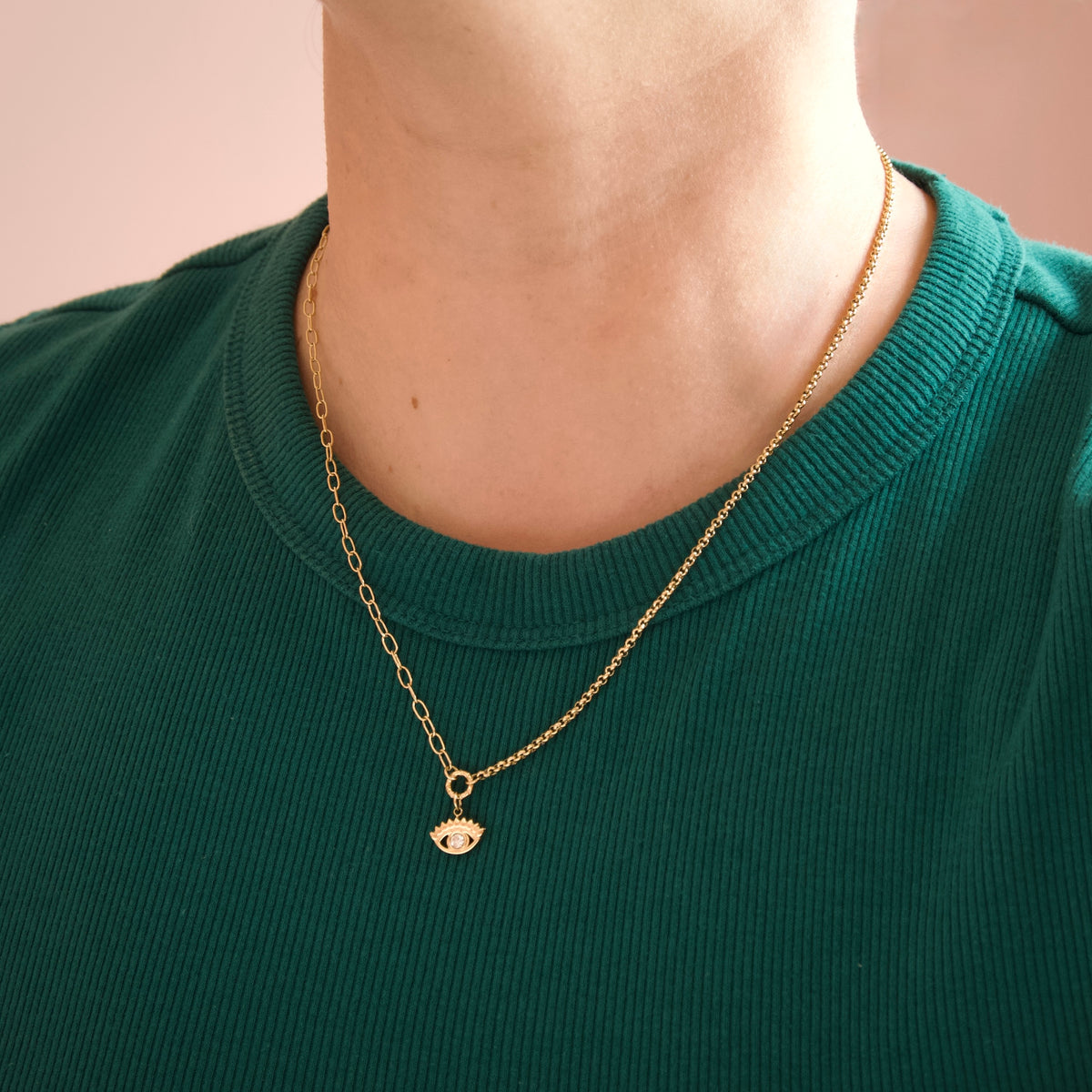 A close-up of a person's neck and upper chest wearing a delicate gold chain necklace with a small evil eye pendant, paired with a dark green ribbed crewneck shirt against a soft pink background.