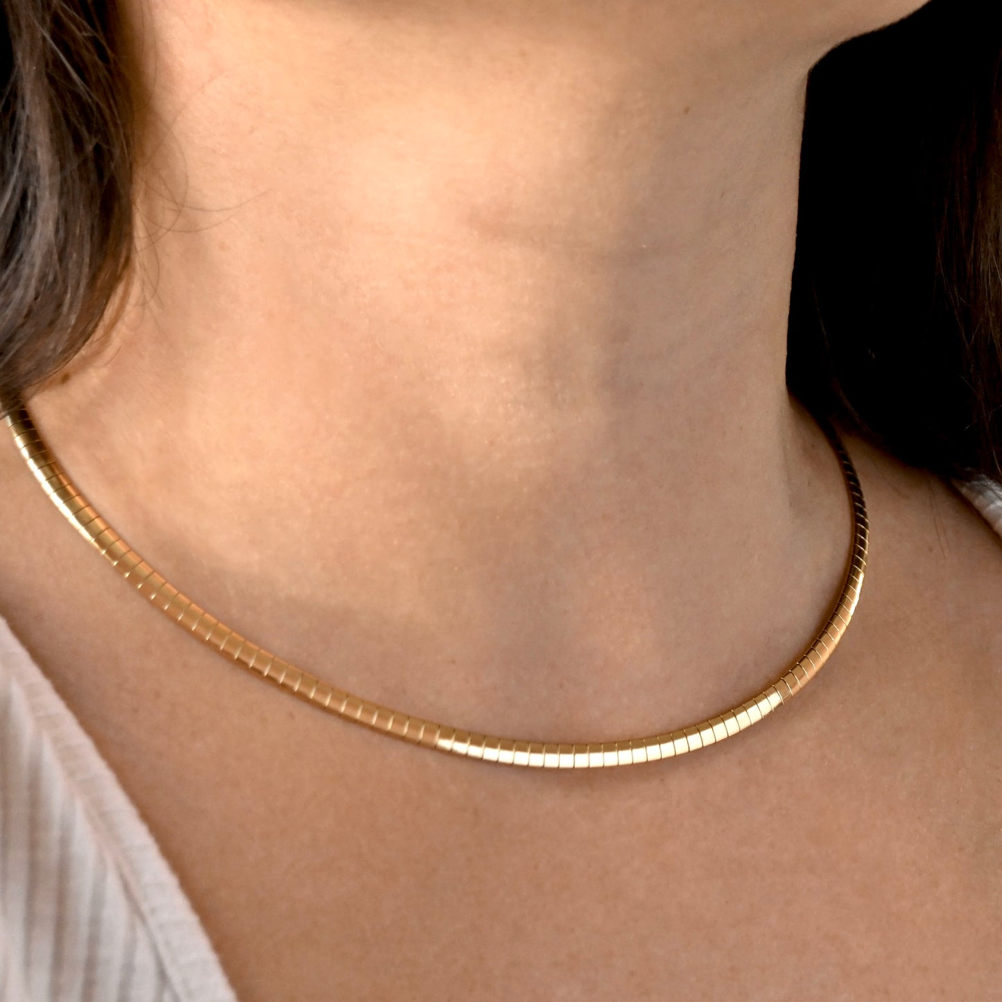 A close-up of a person's neck and collarbone wearing a thin, textured gold chain necklace.