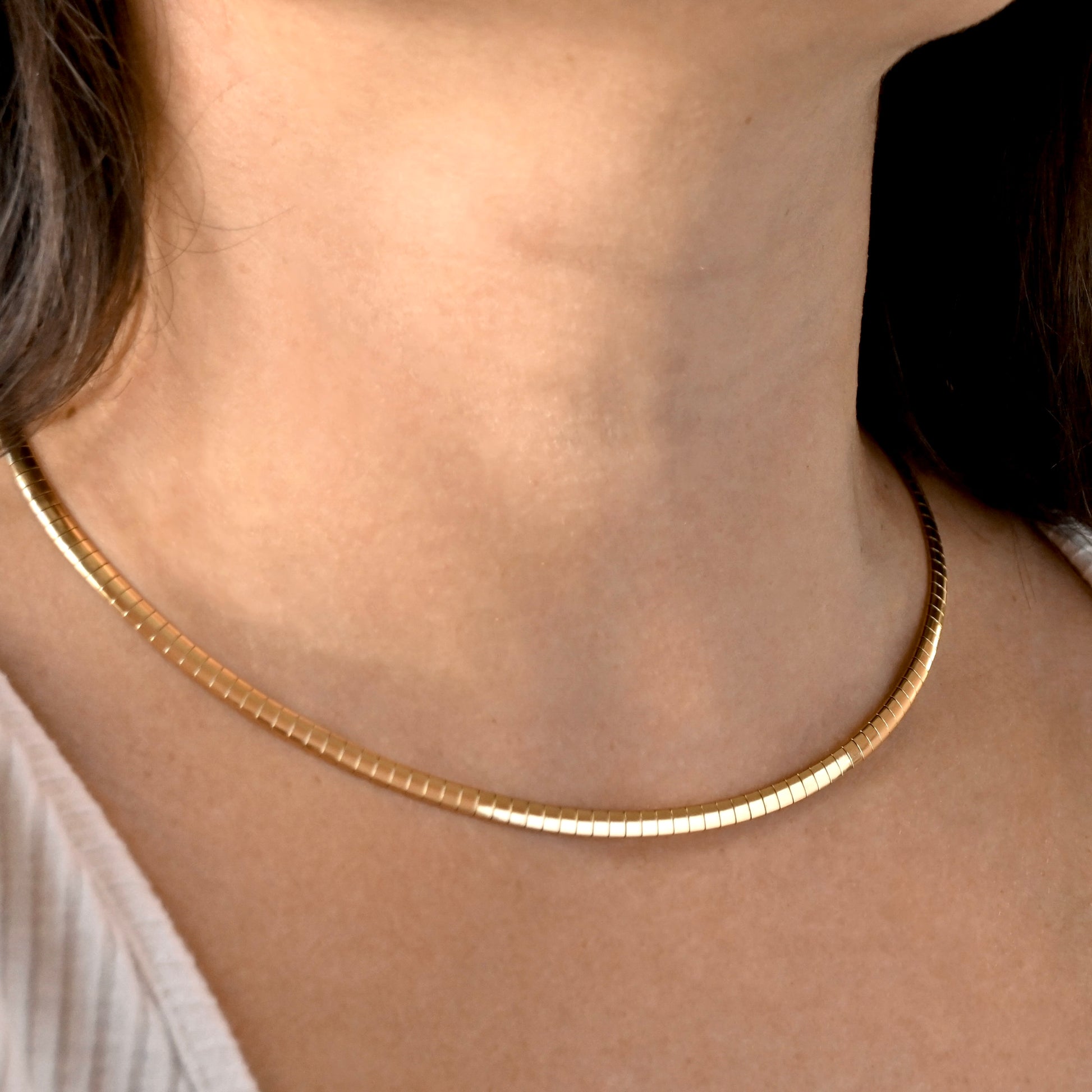 A close-up of a person's neck and collarbone wearing a thin, textured gold chain necklace.