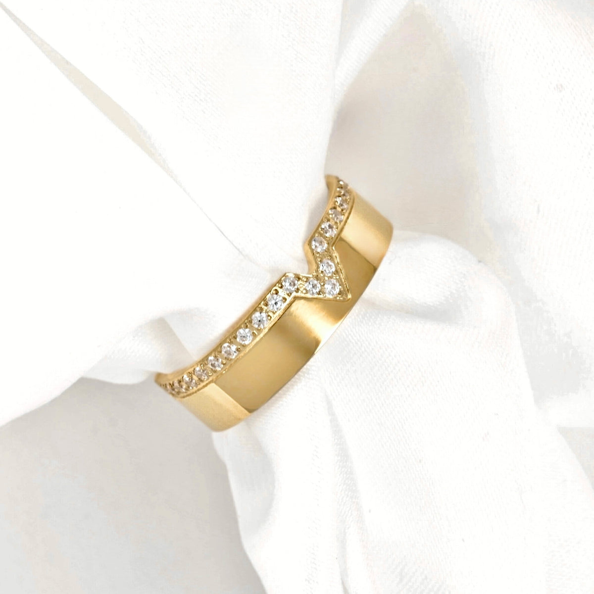 A gold ring with a chevron design, featuring a row of clear pavé stones on one side, displayed on a white fabric napkin.
