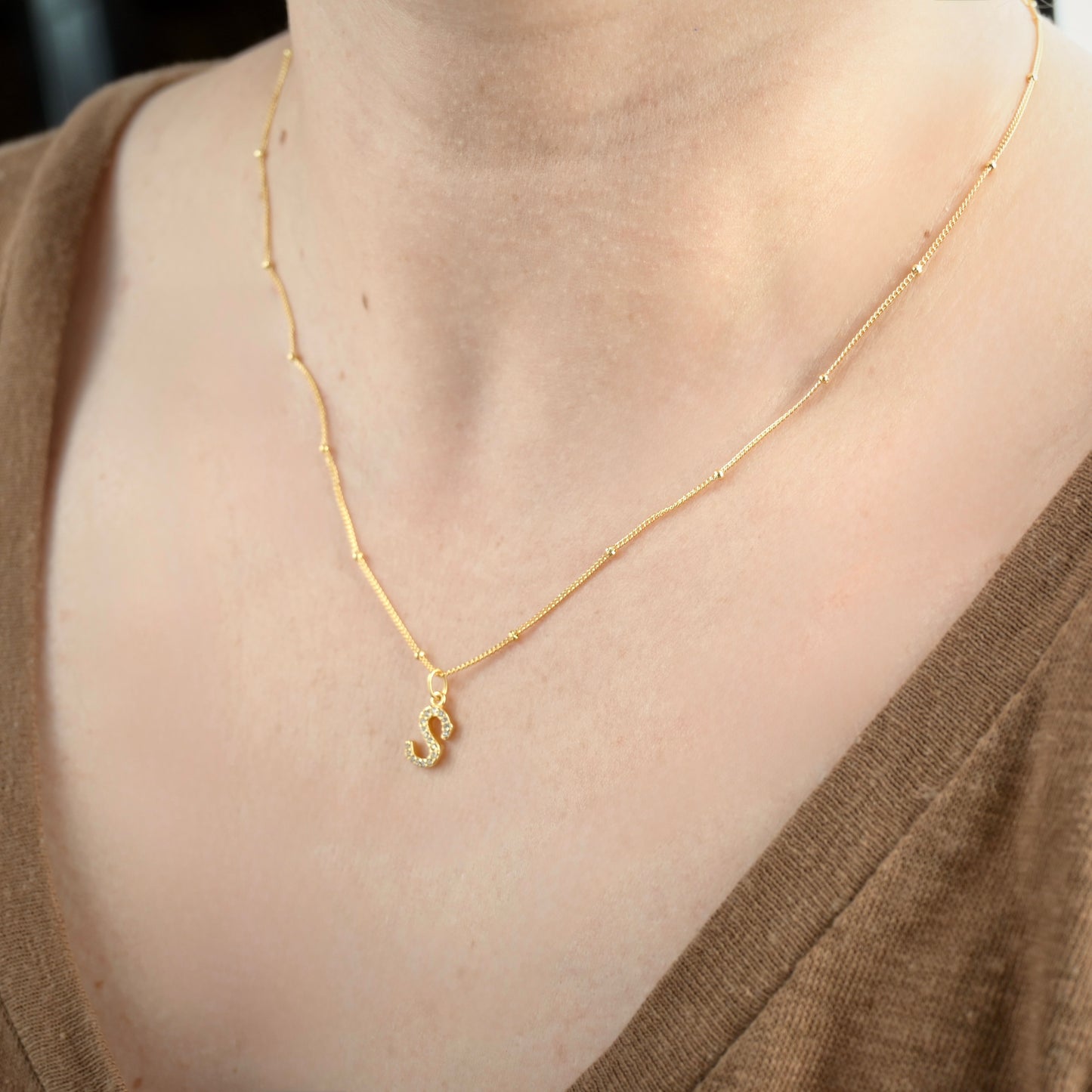 Gold necklace with a small 'S' pendant on a person wearing a brown top.