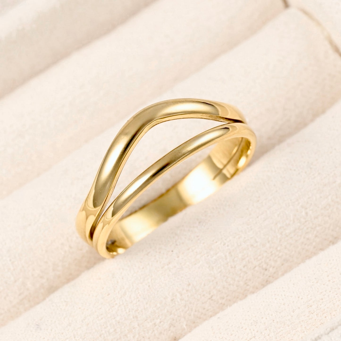 A polished yellow gold ring with a double, flowing wave-like band design, displayed on a textured cream-colored ring cushion.