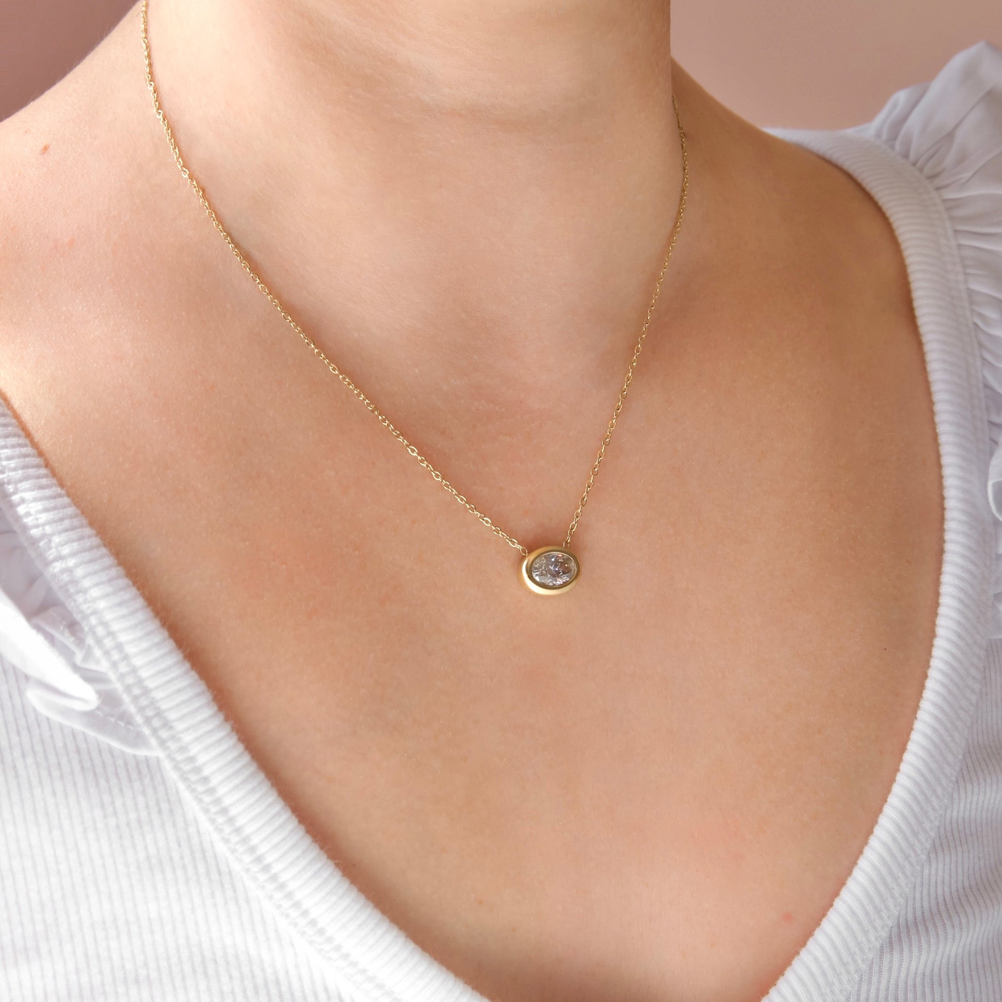 A delicate gold chain necklace with a small oval bezel-set clear gemstone pendant, shown worn on a model's neck over a white ribbed top.