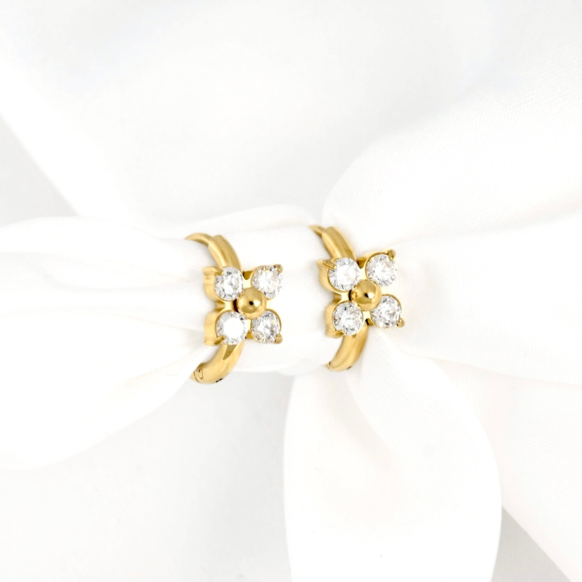 A pair of gold huggie hoop earrings with a flower design set with clear round-cut gemstones, displayed on a white fabric ribbon.
