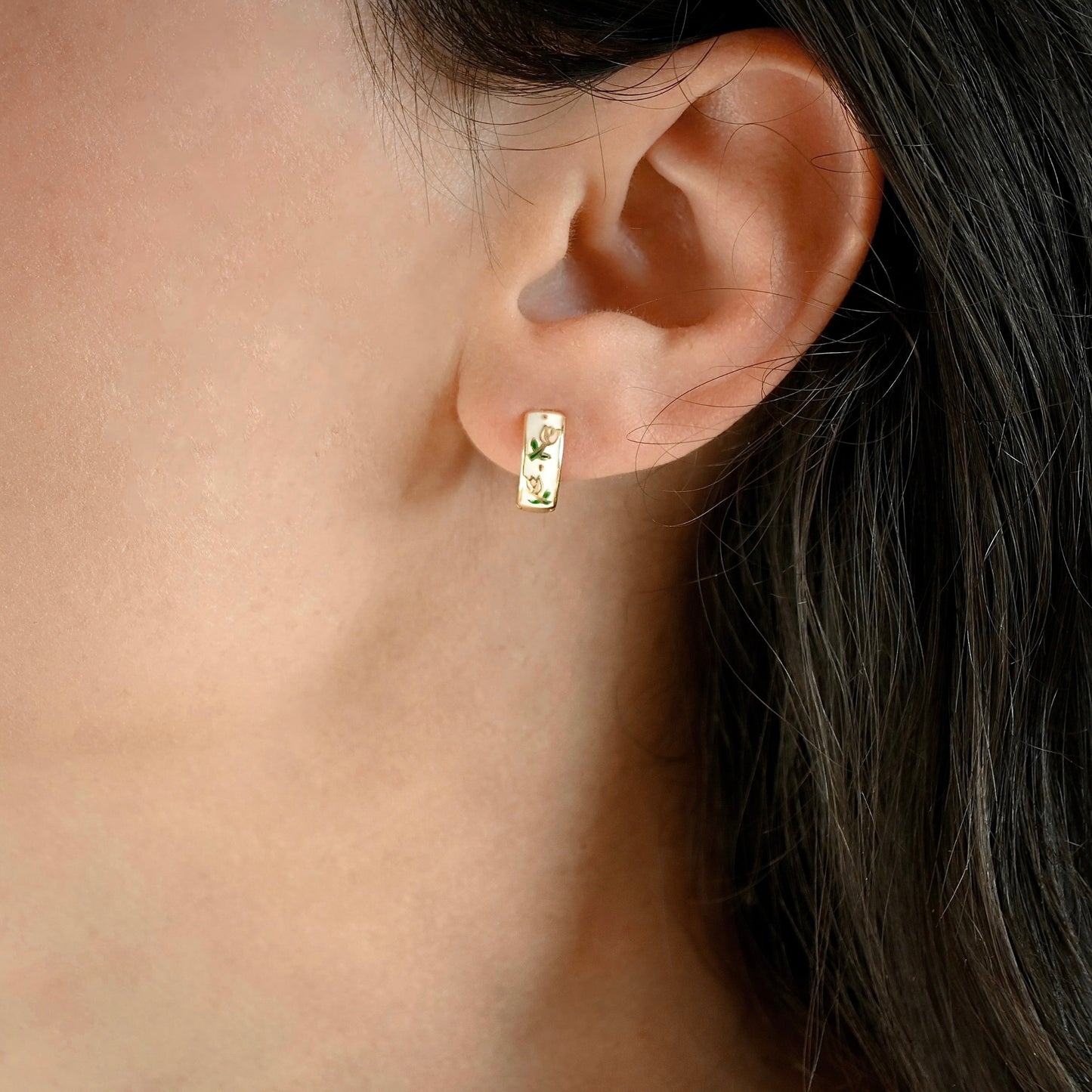 A close-up of a person's ear wearing a small gold hoop earring decorated with delicate green and pink floral details on a white enamel inlay.