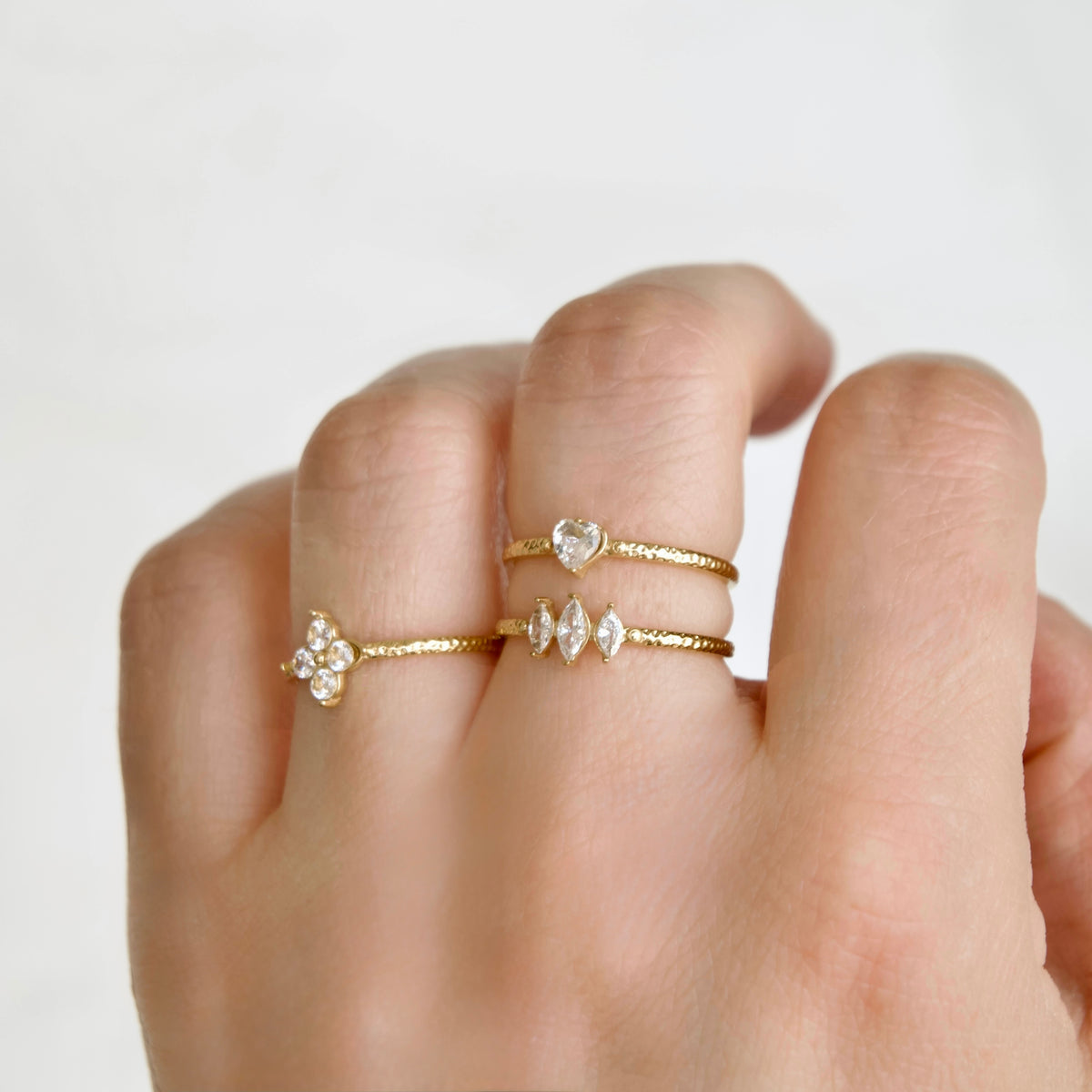 A close-up of a hand wearing three delicate gold rings with clear gemstones, including a heart-shaped stone and marquise-cut stones, against a soft white background.