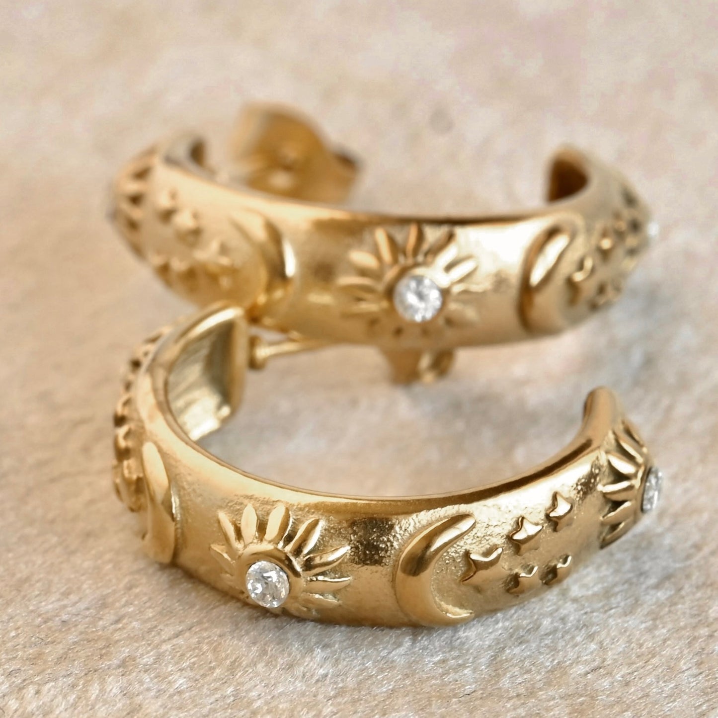 Two gold sun earrings with celestial patterns and small stones on a beige fabric background