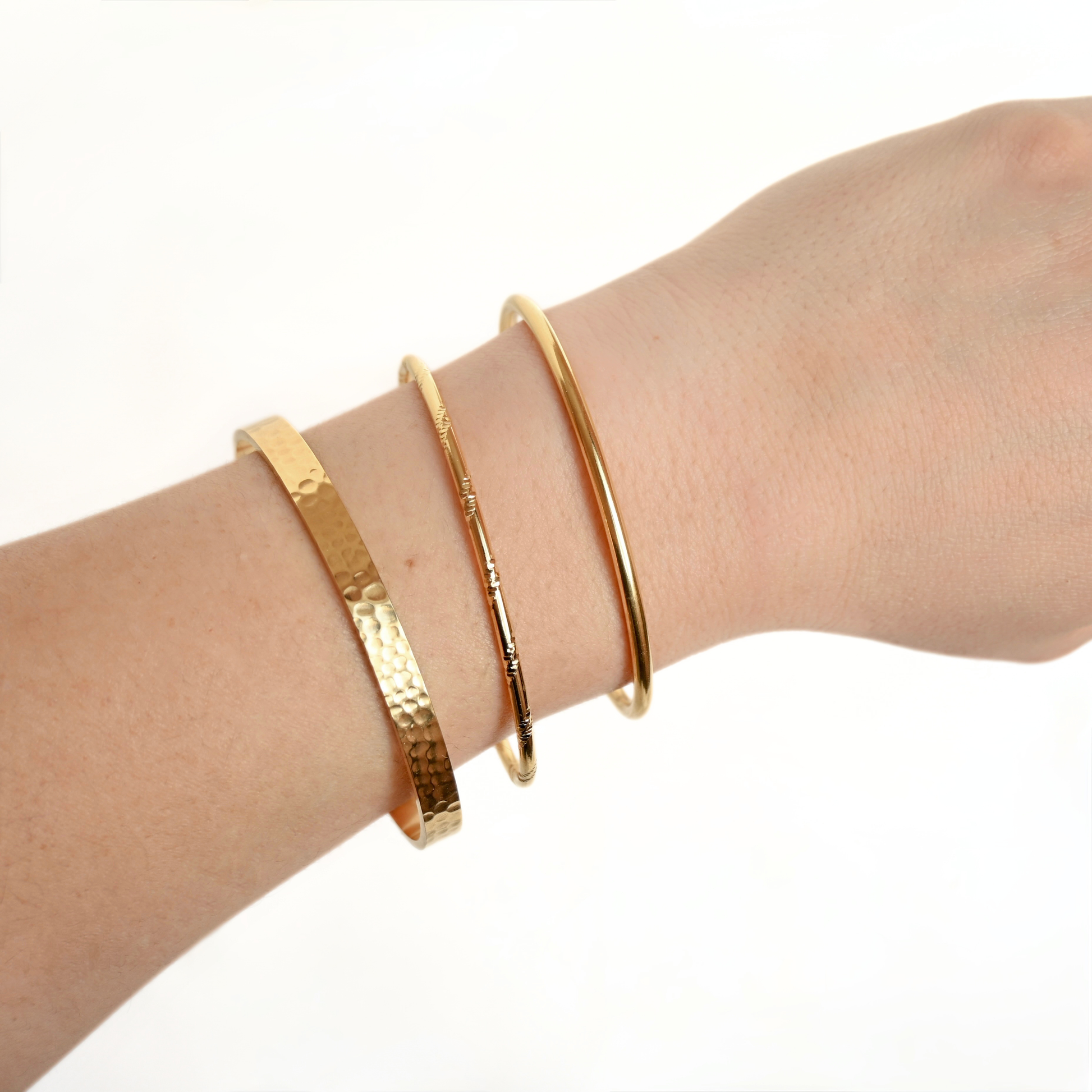 Three gold-toned cuff bracelets worn on a person's wrist, including one wide hammered band, one engraved band, and one smooth slim band, against a plain white background.