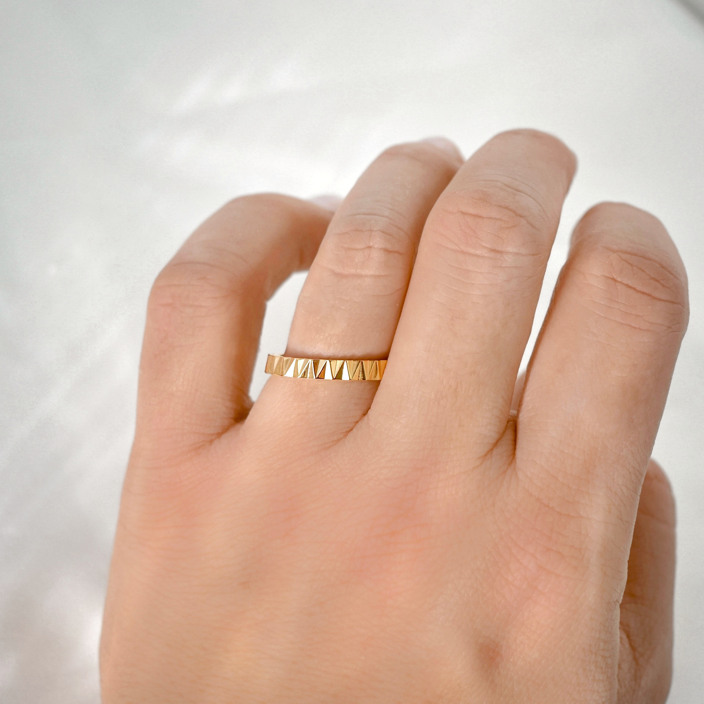 A close-up of a hand wearing a thin gold ring with a textured, geometric triangular pattern on a soft, light gray background.
