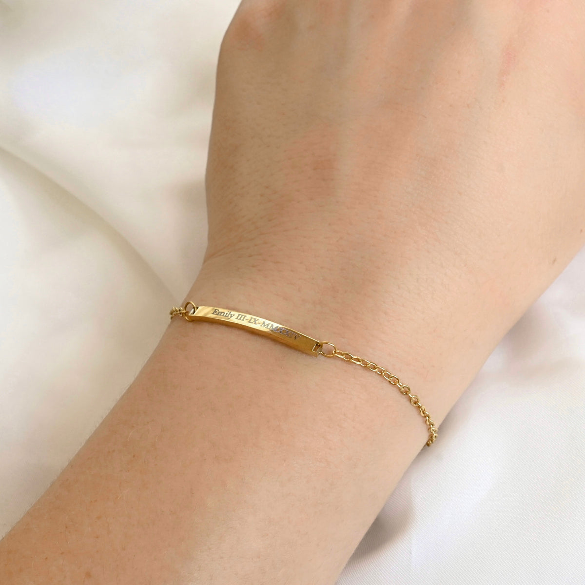 A delicate gold chain bracelet with a polished bar engraved with 'Emily' and a date in roman numerals, worn on a person's wrist against a soft white background.