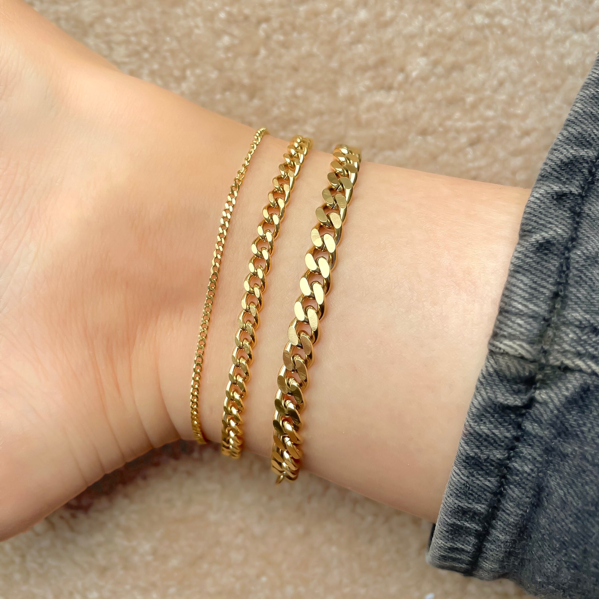 Three gold cuban chain anklets in 2mm, 5mm and 7mm widths stacked on an ankle against a neutral background 