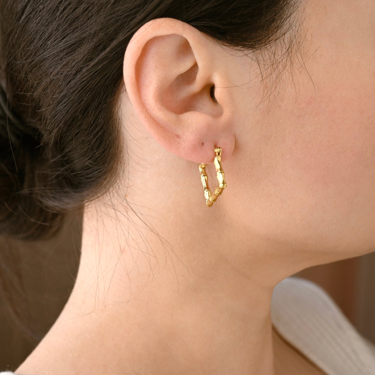 A close-up of a model's ear wearing a small textured square gold hoop earring.