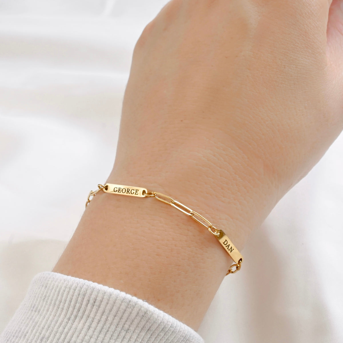 A close-up of a person's wrist wearing a gold paperclip chain bracelet with engraved names 'GEORGE' and 'DAN', paired with a white ribbed knit sleeve against a soft white background.