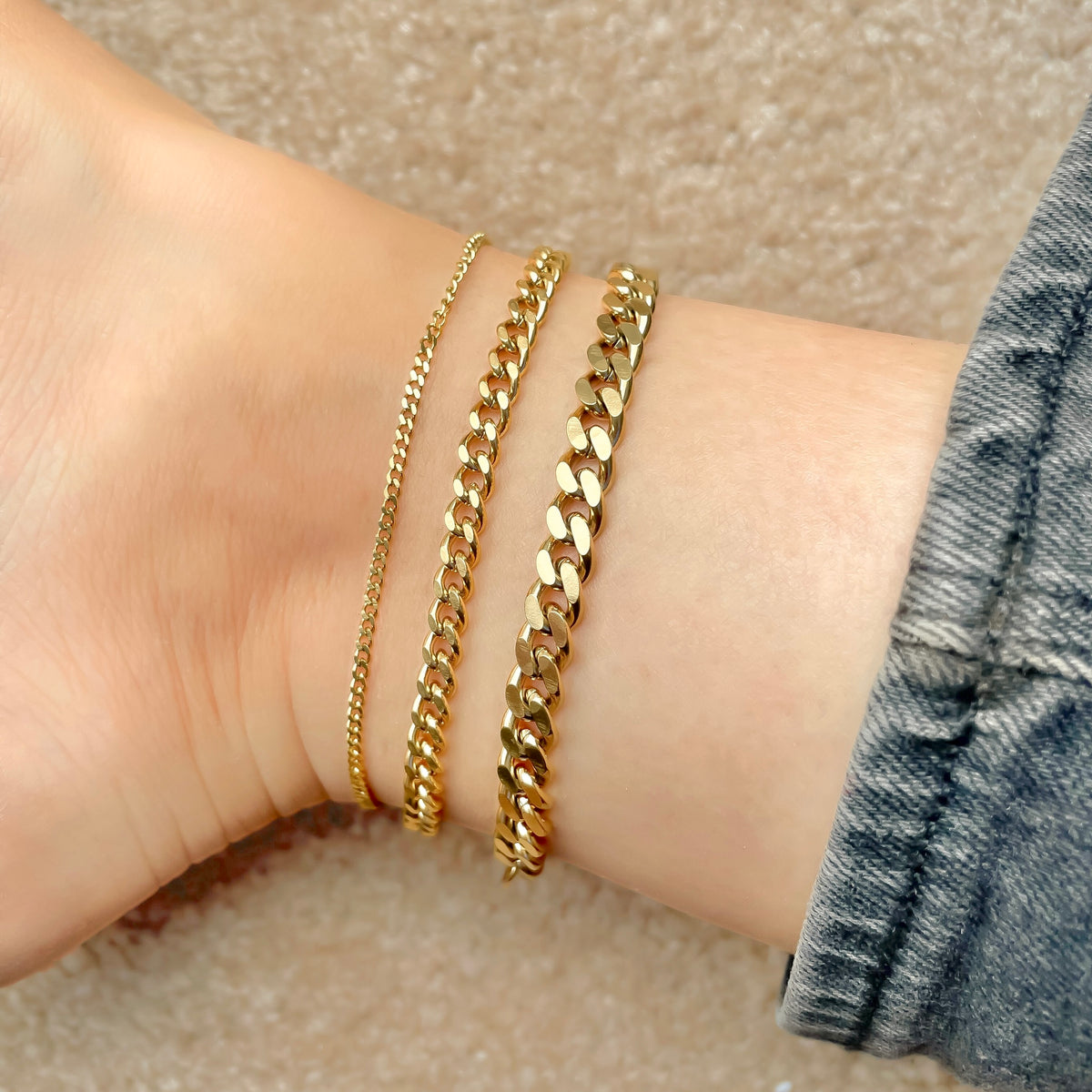 Three gold cuban chain anklets in 2mm, 5mm and 7mm widths stacked on an ankle against a neutral background 