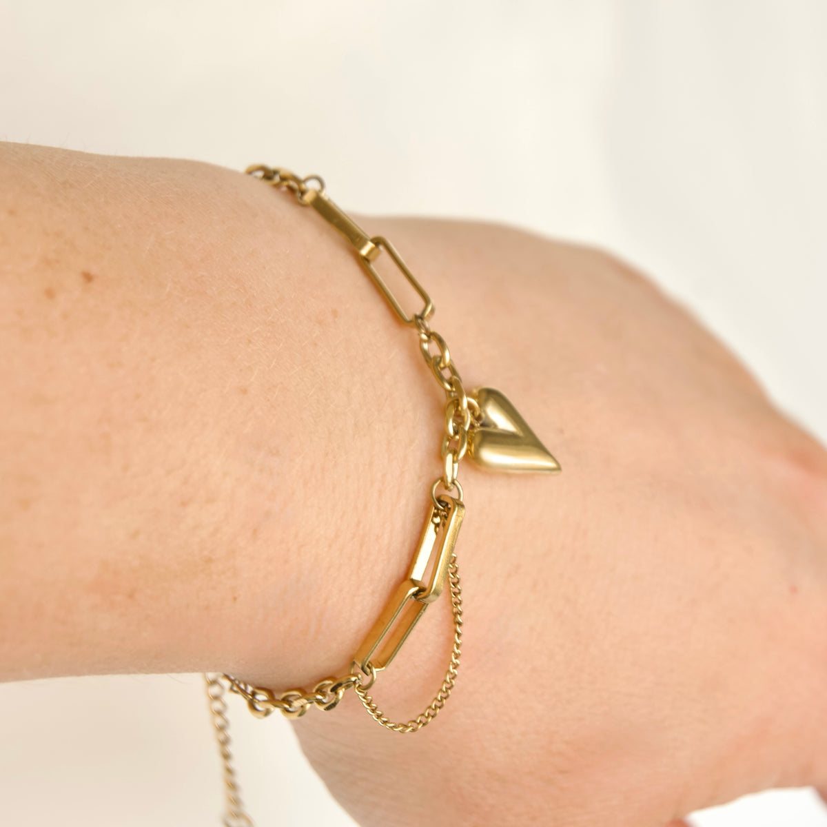 A gold chain bracelet with rectangular links, a small heart charm, and a safety chain, shown worn on a person's wrist against a plain light background.