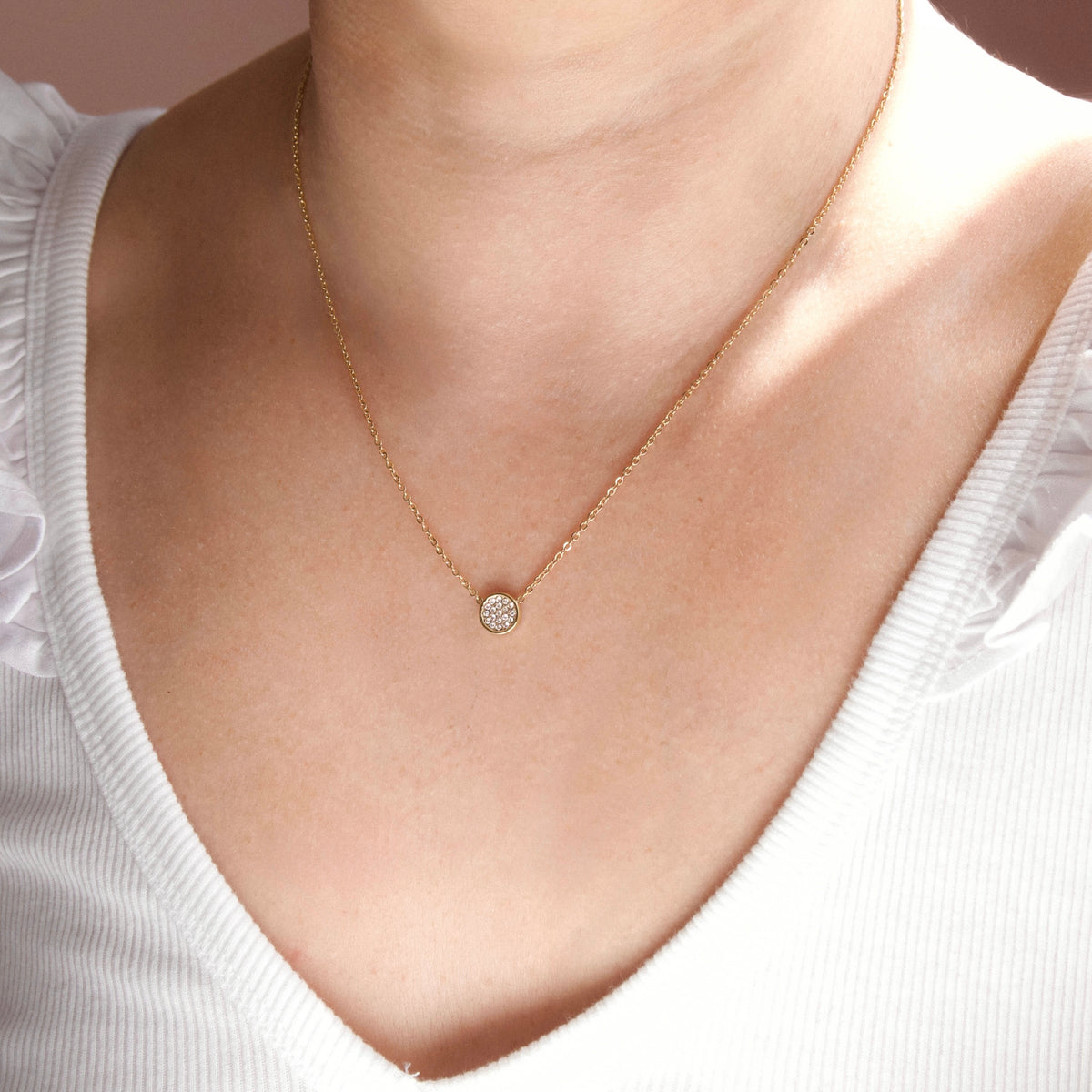 A delicate gold chain necklace with a small round, clear gemstone pendant, worn by a model in a white ribbed top.