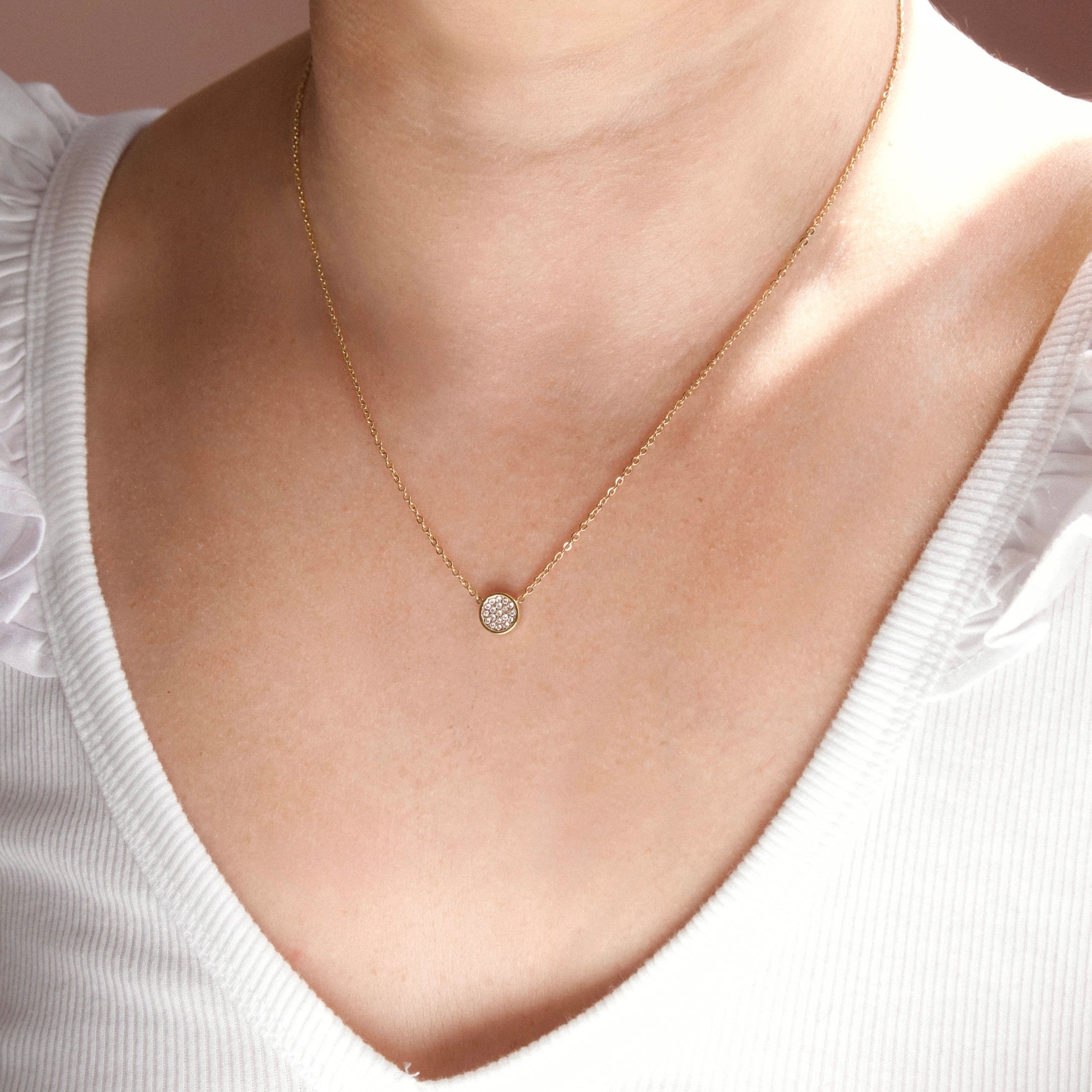 A delicate gold chain necklace with a small round, clear gemstone pendant, worn by a model in a white ribbed top.