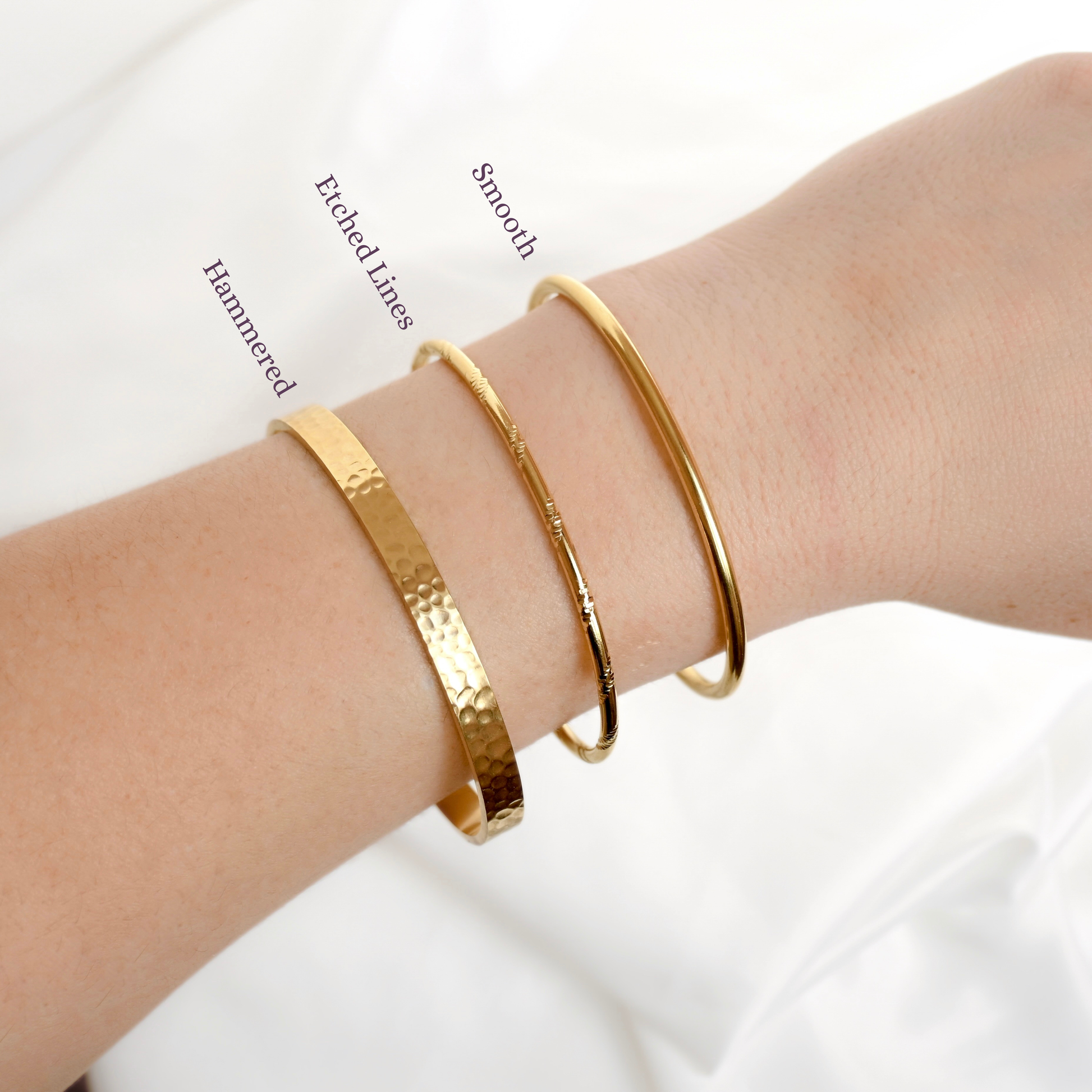 Three gold bangle bracelets with different textures (hammered, etched lines, smooth) shown on a person's wrist against a white fabric background.