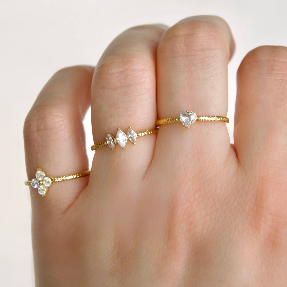 Three delicate gold rings with clear gemstones worn on a hand. One ring features a small four-petal flower cluster, while the other two have textured gold bands with marquise and heart-shaped clear stones.